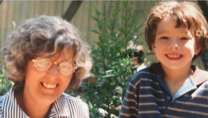 A woman with curly gray hair and glasses smiling next to a young boy with curly hair and a striped shirt, outdoors with greenery in the background.