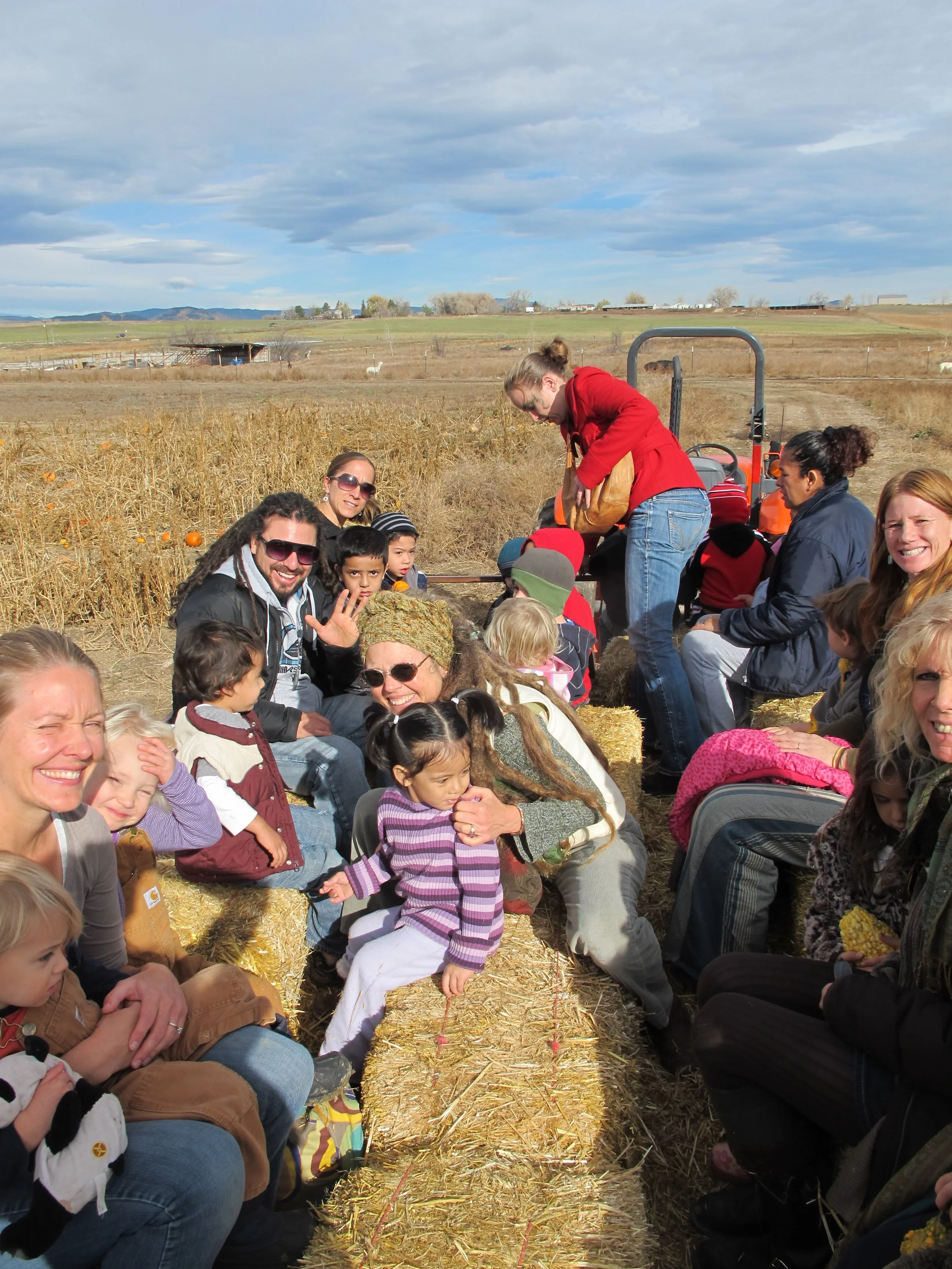 Group of people, including children and adults, sitting and standing on hay bales in an open field during daytime. Some are smiling, and a woman in a red jacket is leaning over a tractor in the background. The field has a straw pathway, and there are hills, farm buildings, and a partly cloudy sky in the distance.