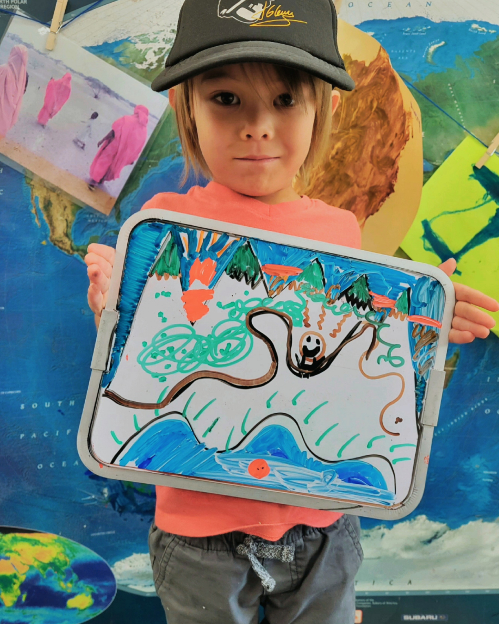 Child holding a whiteboard with a colorful drawing of mountains, trees, and a sun. Background features a world map and an ocean map.