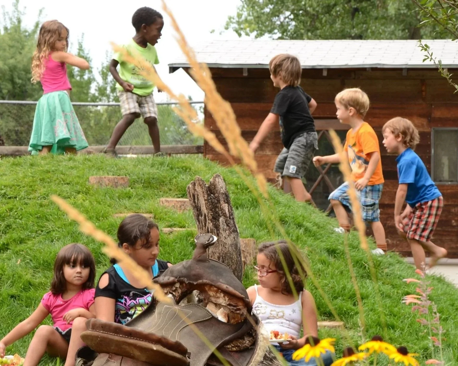 Children gathered outside on a grassy hill, some sitting and some standing, with tall grass and a wooden building in the background.