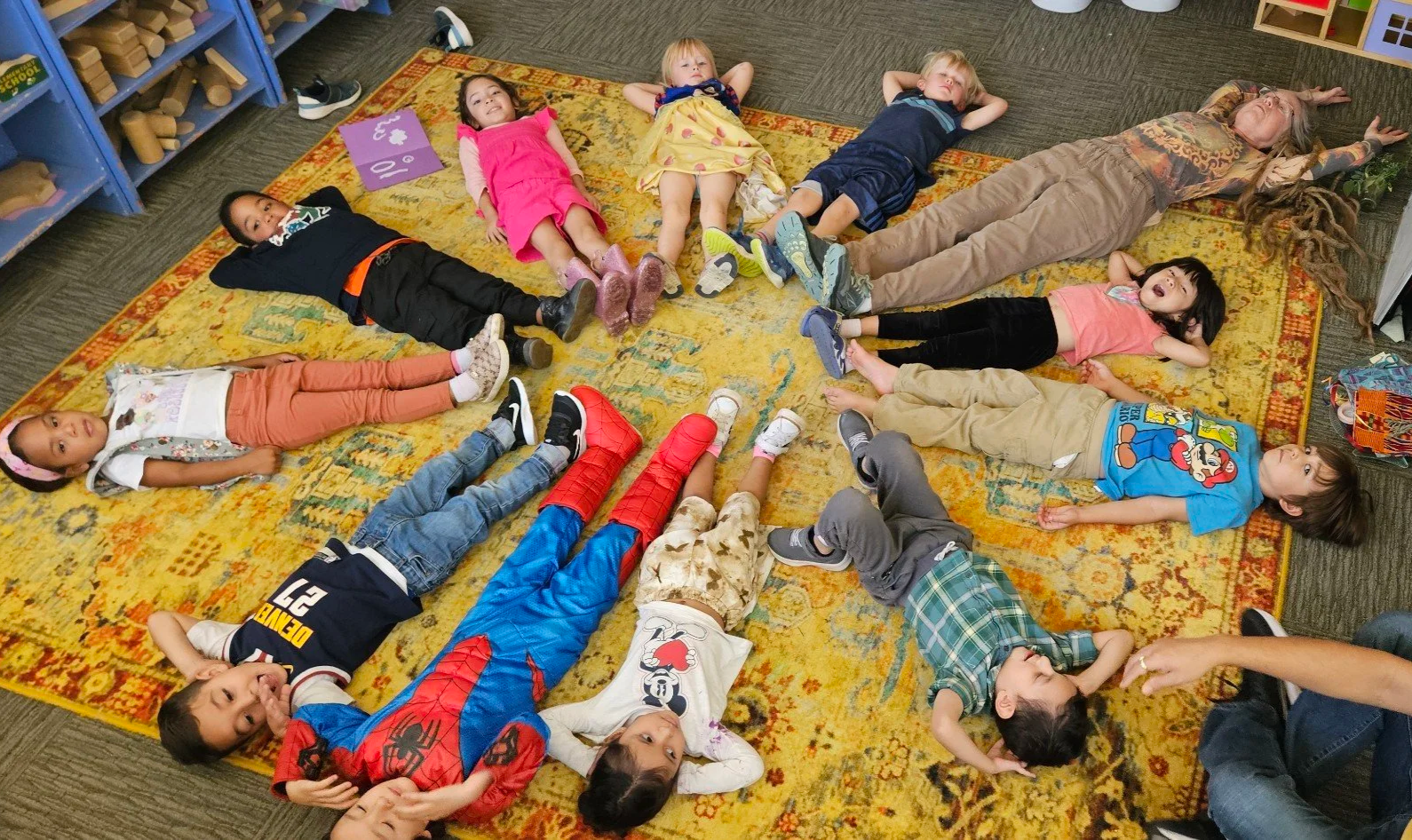 Children lying on a colorful rug in a circle with an adult at the edge, in a classroom setting.