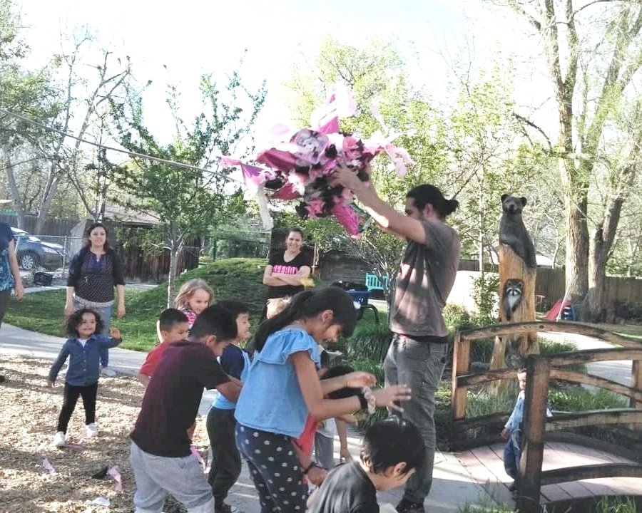 A group of children and adults outdoors, with a man holding a pinata, and children reaching out to grab or touch them, in a park-like setting with trees and a wooden fence.