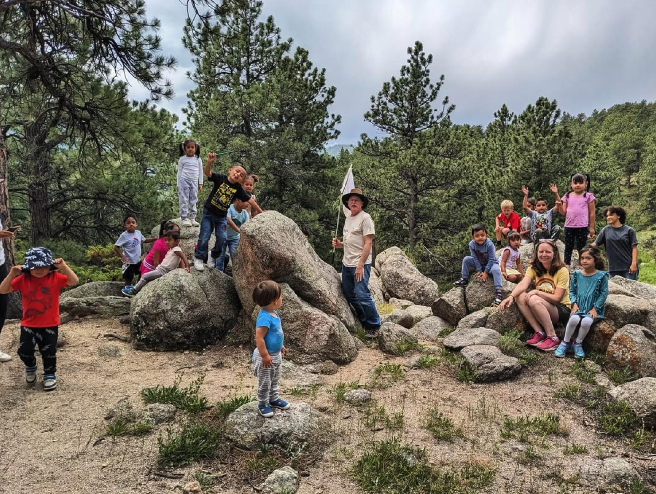 Children and adults gathered outdoors in a forested area with large rocks, some children standing on rocks and others sitting or standing nearby, under a cloudy sky.
