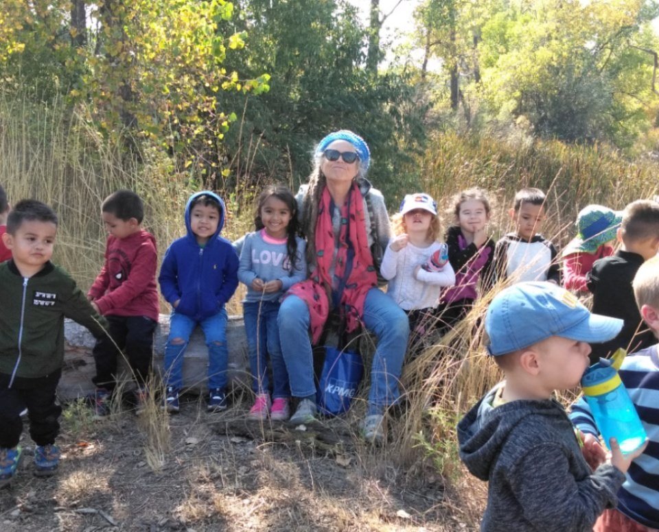 A woman surrounded by young children outdoors on a nature hike, some sitting on a fallen log, others standing, with trees and tall grass in the background.