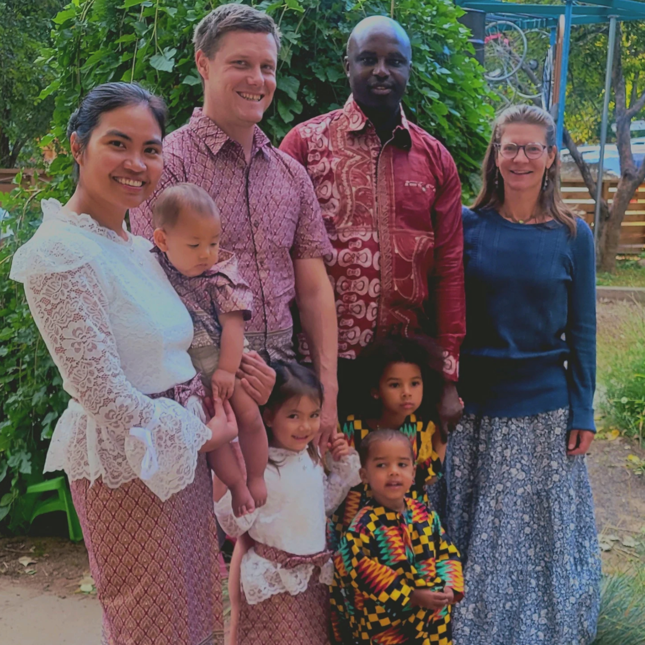 Group of six adults and four children outdoors in a garden, with green foliage and a wooden fence in the background.