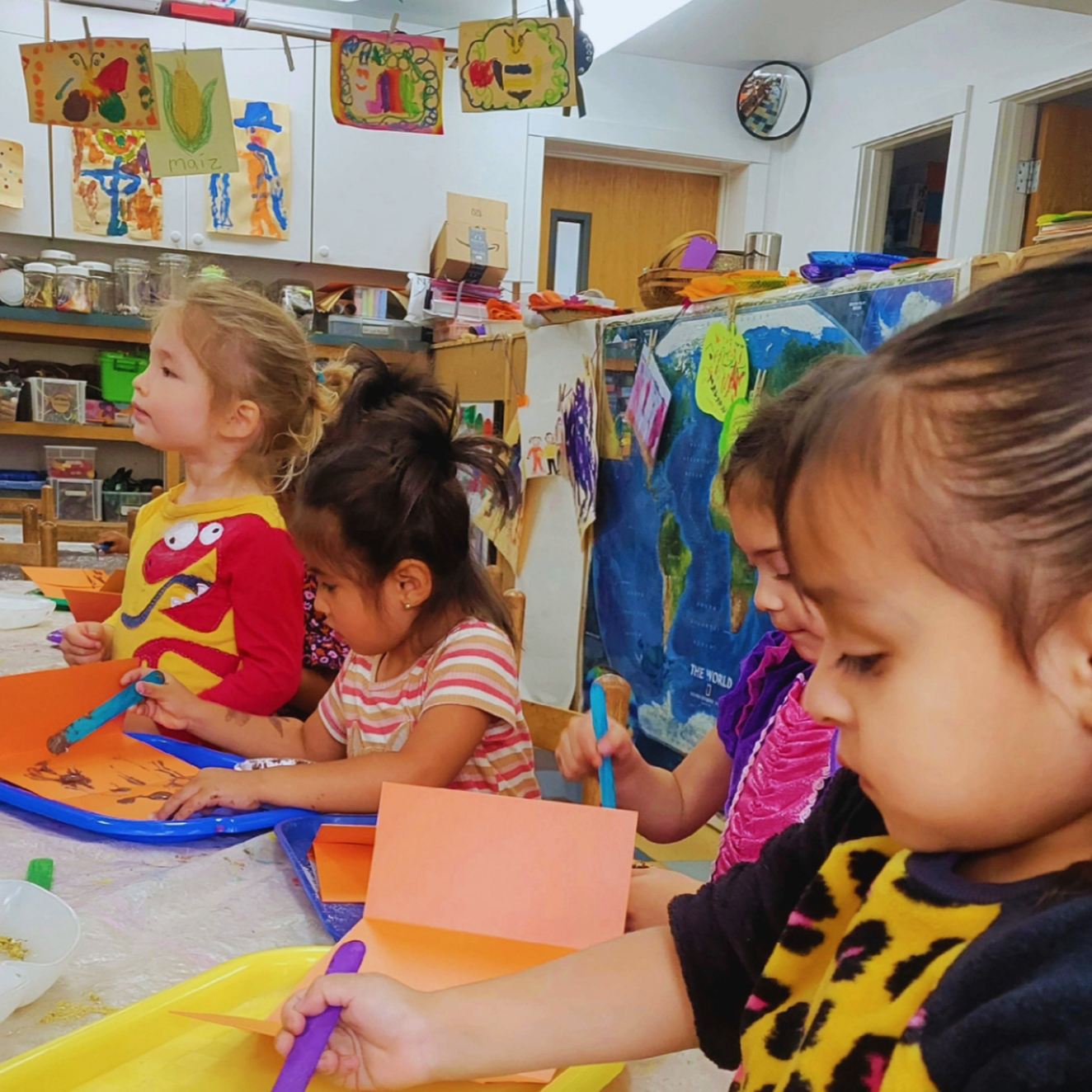 Children sitting at a table engaged in arts and crafts activities in an art classroom, with artwork hanging on the walls and a world map in the background.