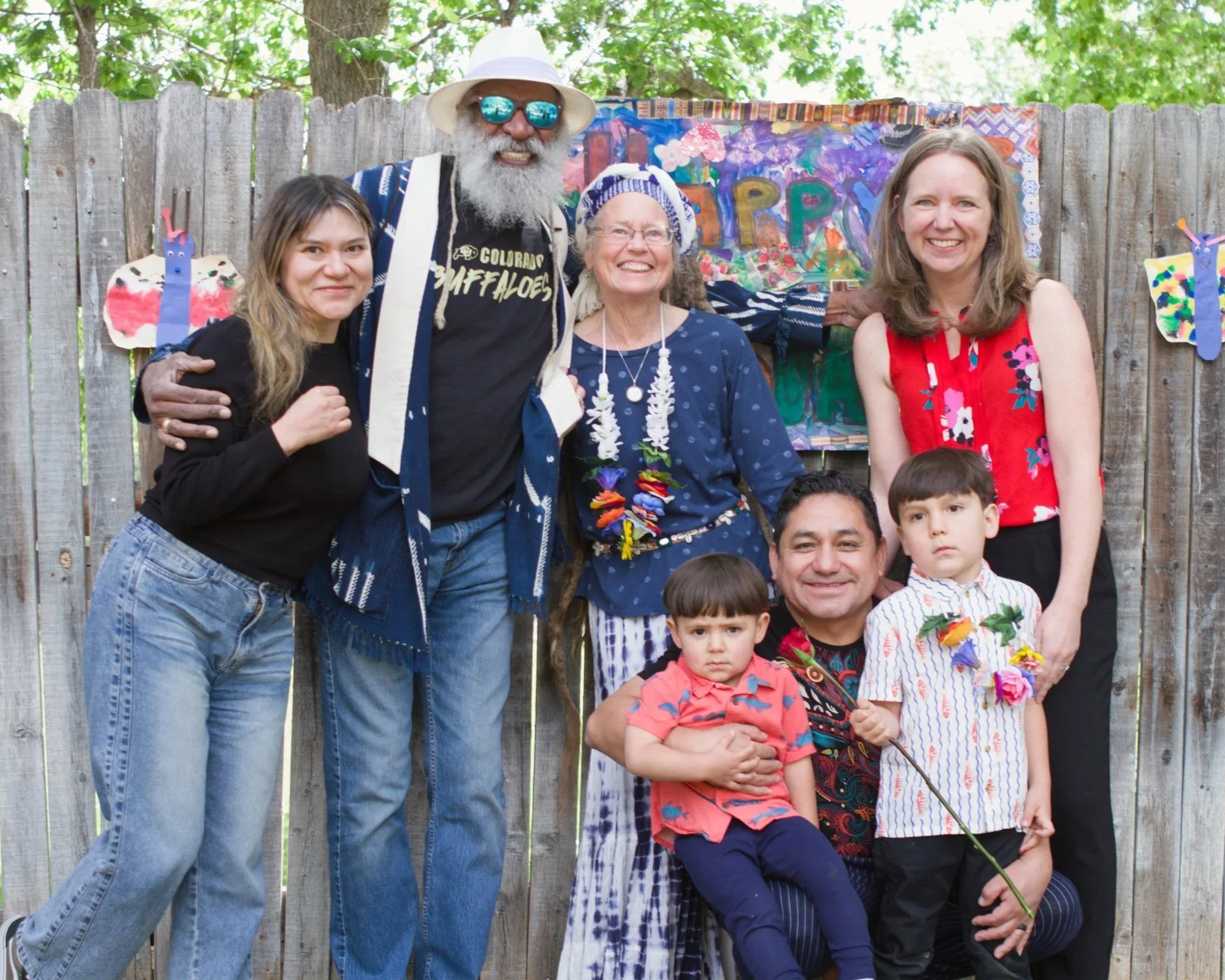 Group of five people smiling in front of a colorful, abstract painted backdrop on a wooden fence, with two children at the front and three adults behind, outdoors with green trees overhead.
