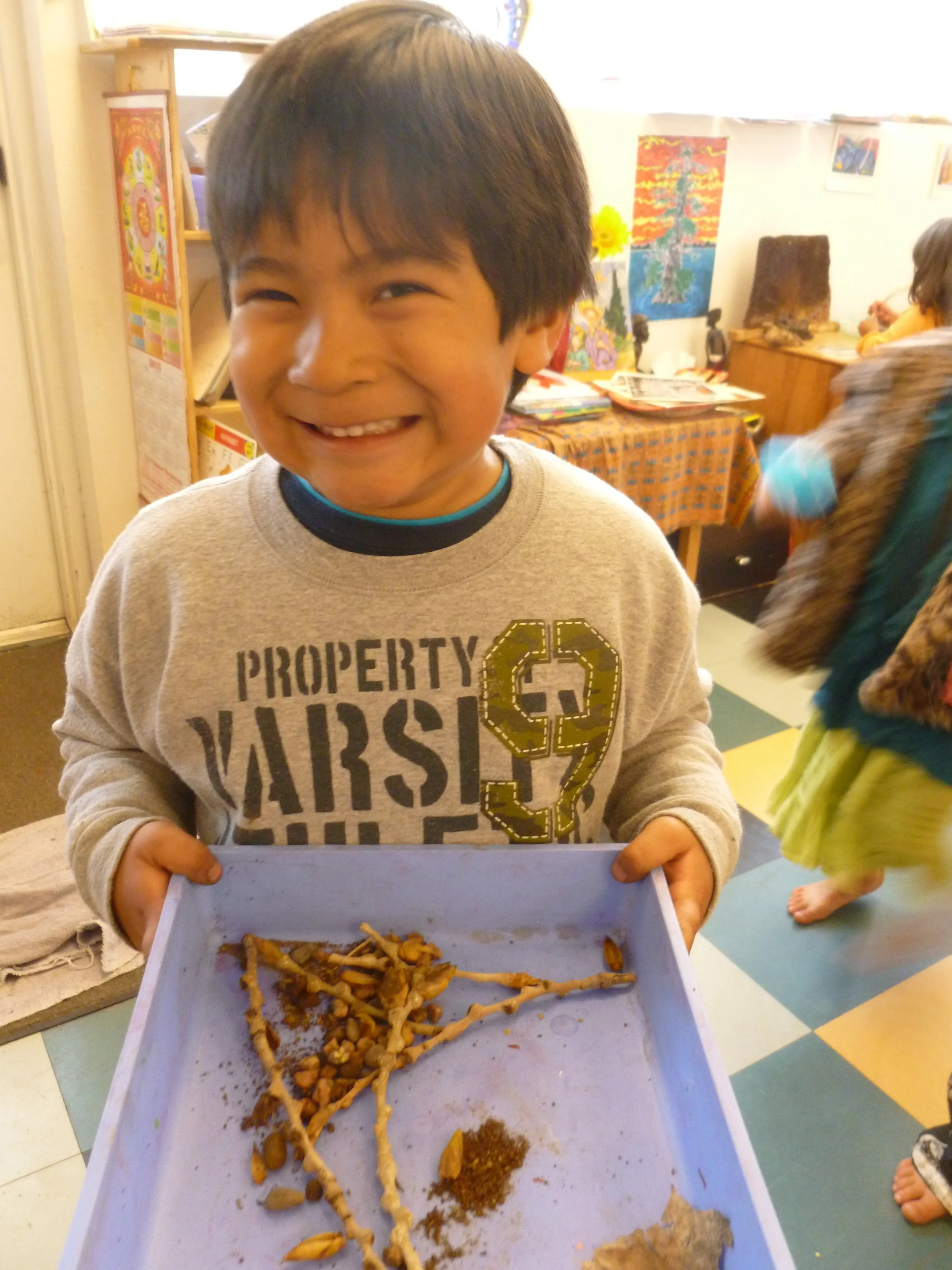 A smiling young boy holding a tray with animal bones and artifacts in an indoor setting with colorful wall art and another child in the background.