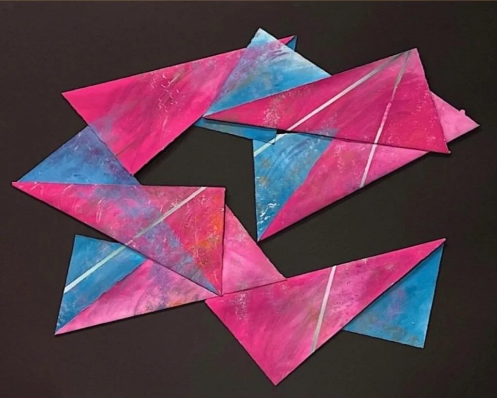 Colorful, painted paper triangles with pink, blue, and white streaks arranged in a circular pattern on a black background.