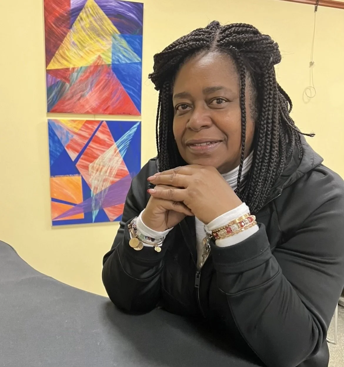A woman with braided hair sitting at a table, resting her chin on her hands, wearing a black jacket and jewelry, with colorful abstract artwork in the background.