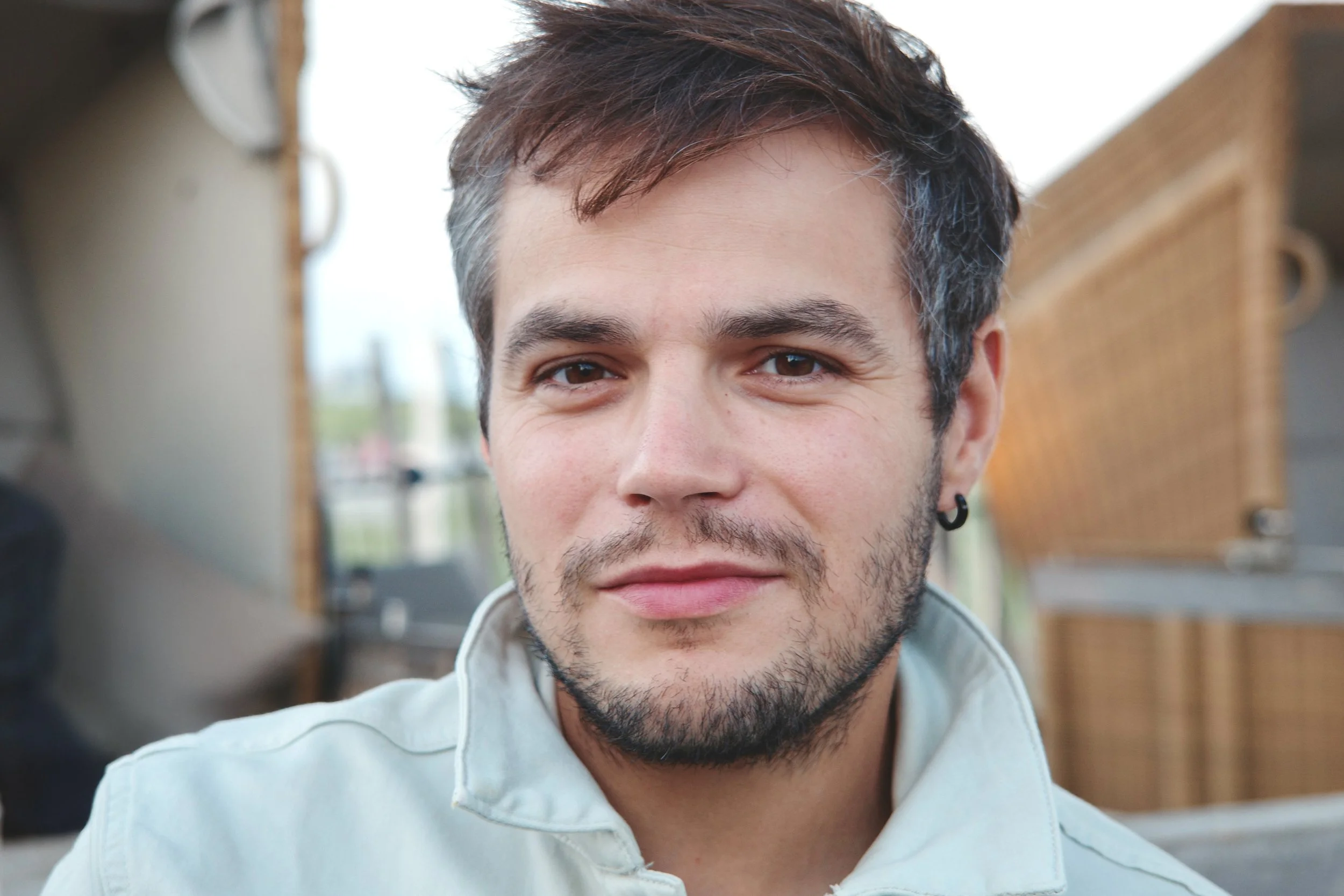 Nahaufnahme eines jungen Mannes mit kurz braunen Haaren, leichtem Bart und Ohrpiercing, der in einem hellen Jacke vor einem Restaurant oder Café steht.