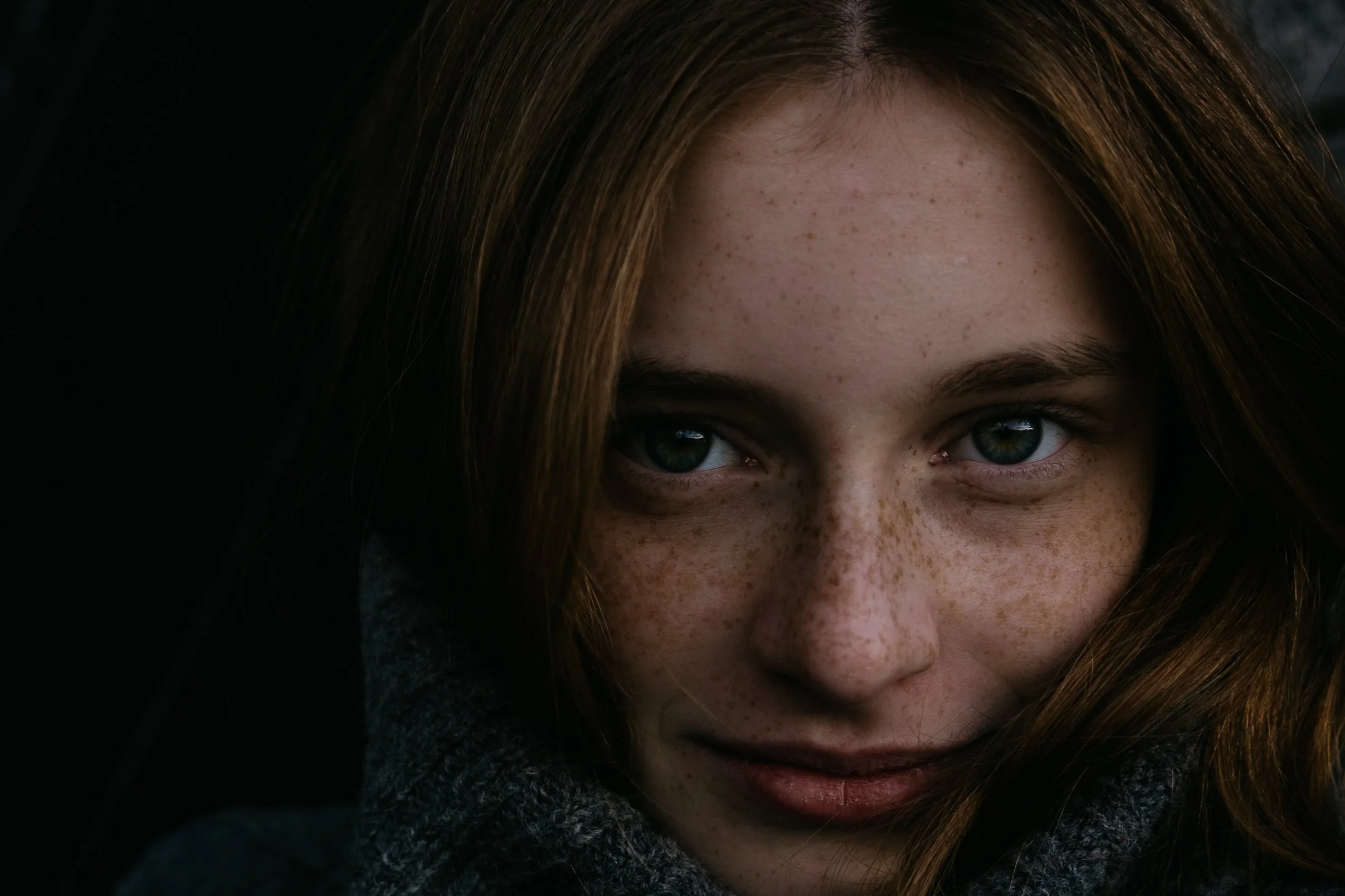Close-up of a young woman with red hair and freckles, smiling slightly, with bright blue eyes and her face partly shadowed.