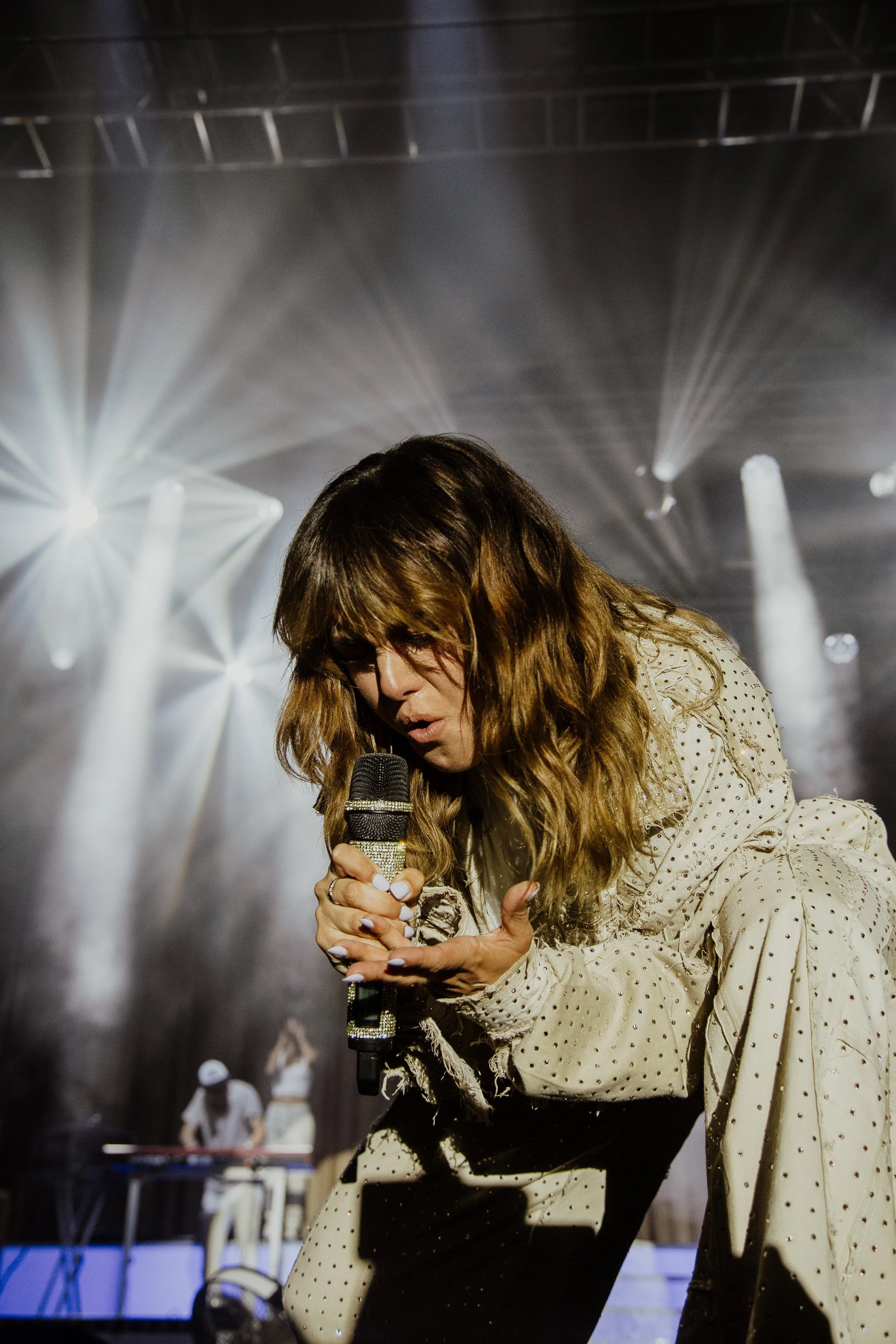 A female singer with long, wavy brown hair, wearing a beige polka-dot outfit, passionately performing on stage, holding a microphone, with stage lights shining in the background and two other musicians in the background.