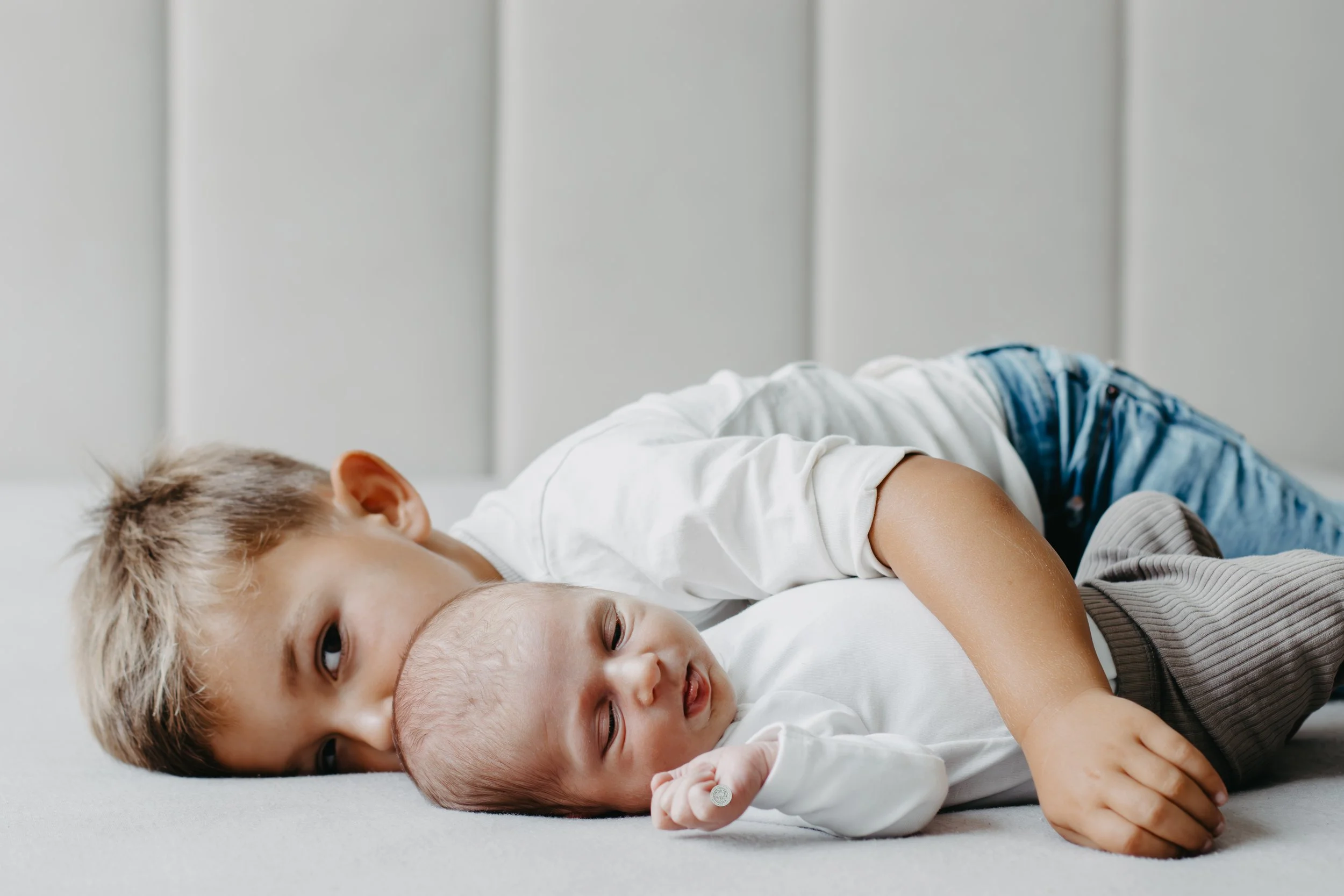 A young boy is lying on the bed, hugging a newborn baby while resting their heads on a white surface.
