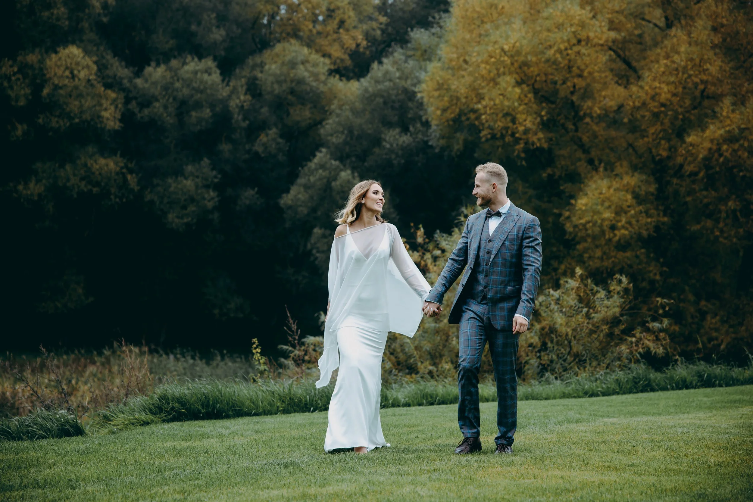 A bride and groom walking hand in hand on a lush green lawn outdoors, with colorful autumn trees in the background, smiling at each other.