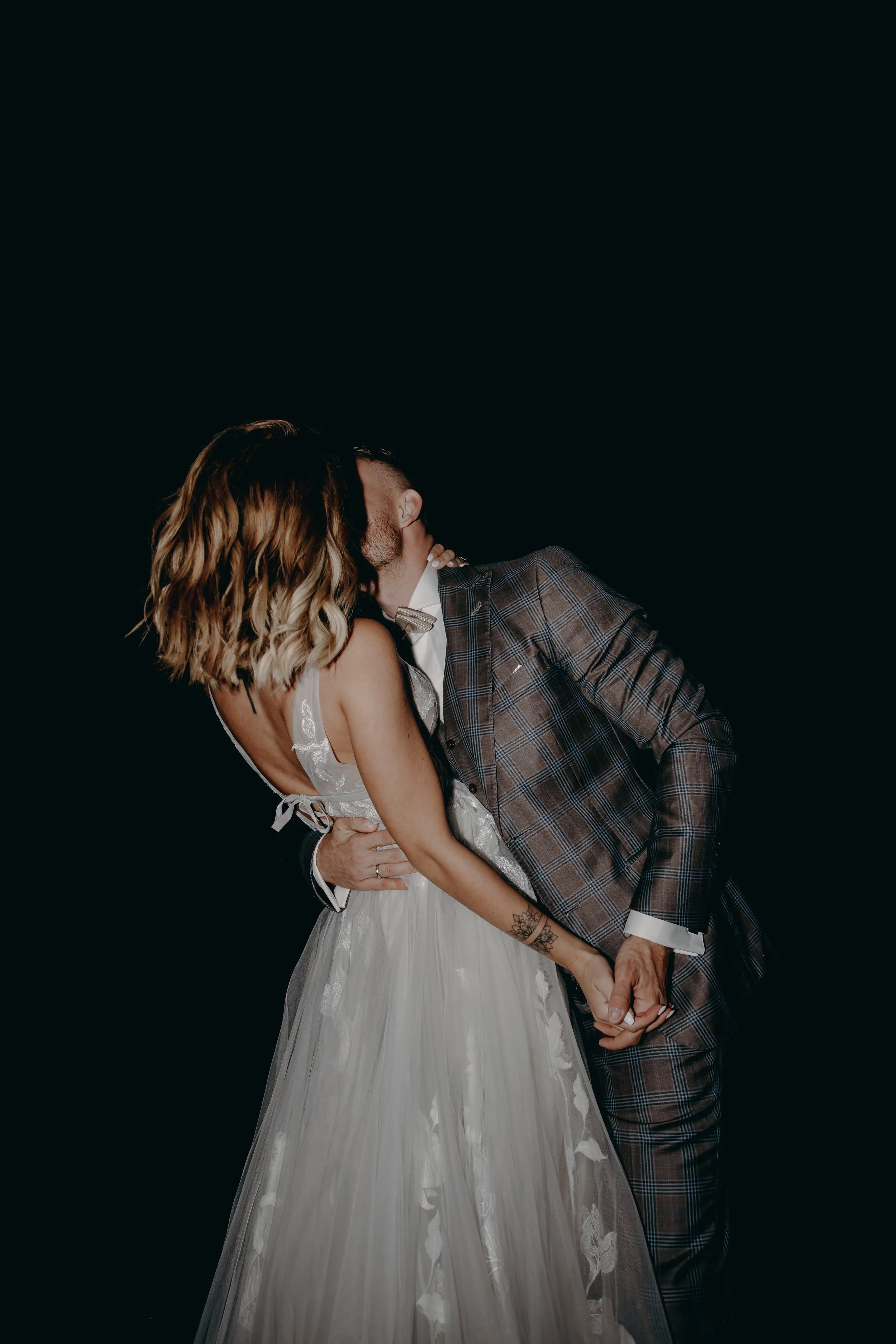 A couple dressed in wedding attire sharing a kiss against a black background, with the bride holding the groom's hand.