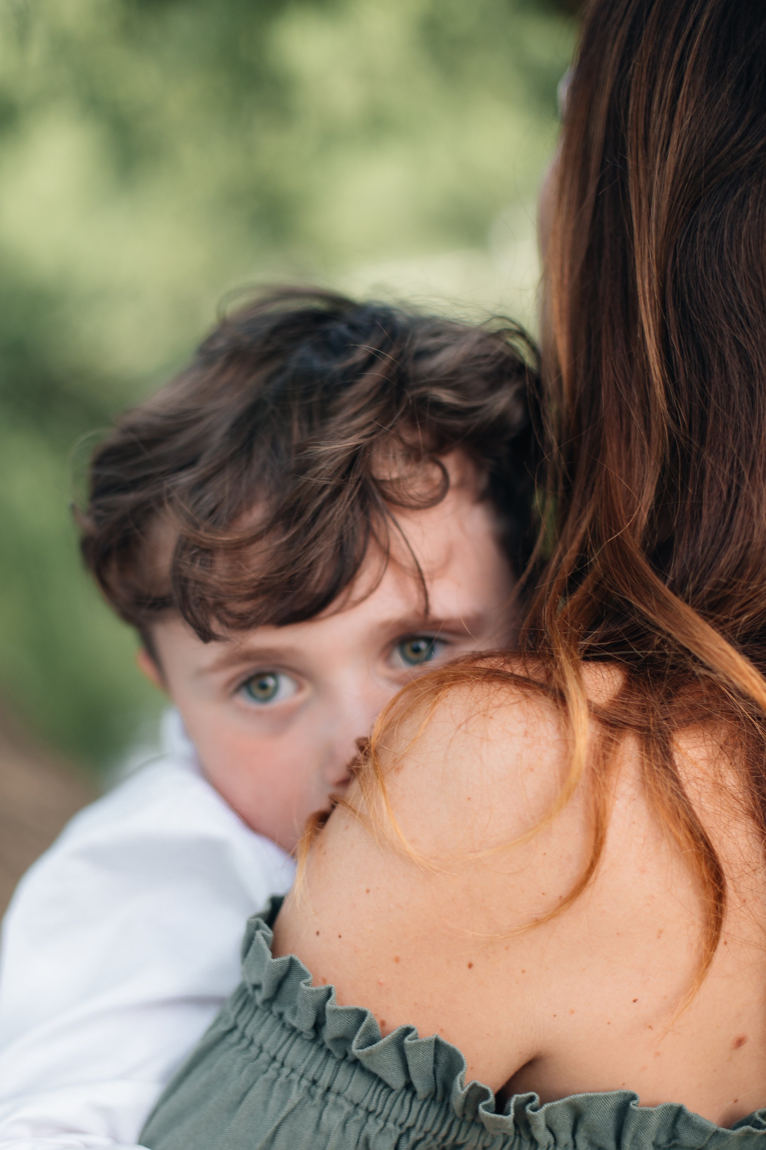 Child with brown curly hair and blue eyes resting on woman with red hair outdoors.
