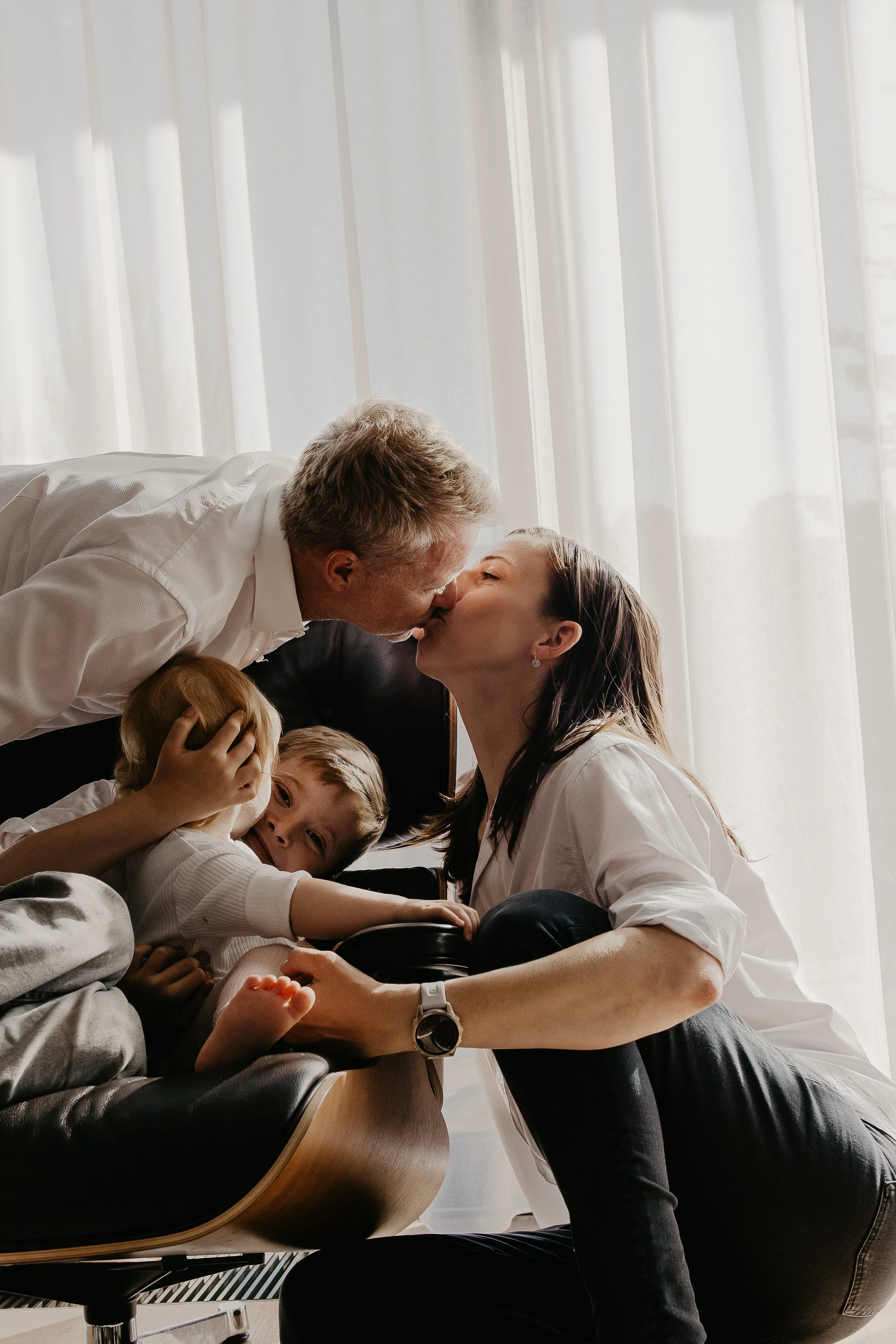 A family sharing a kiss in a cozy living room with natural light coming through sheer curtains.
