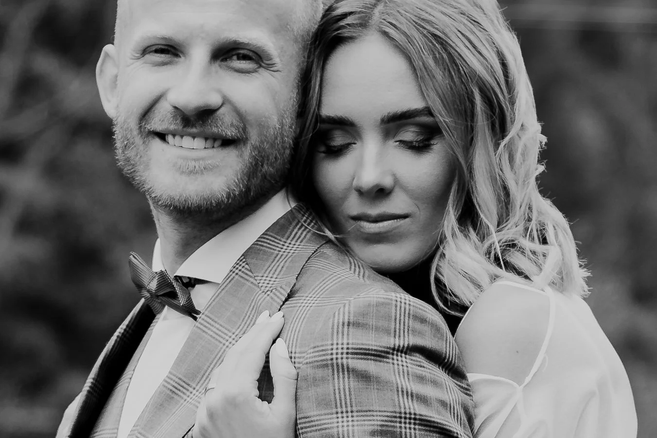 A black-and-white photo of a happy man with a beard and a woman with curly hair, both dressed nicely, smiling and holding each other closely outdoors.