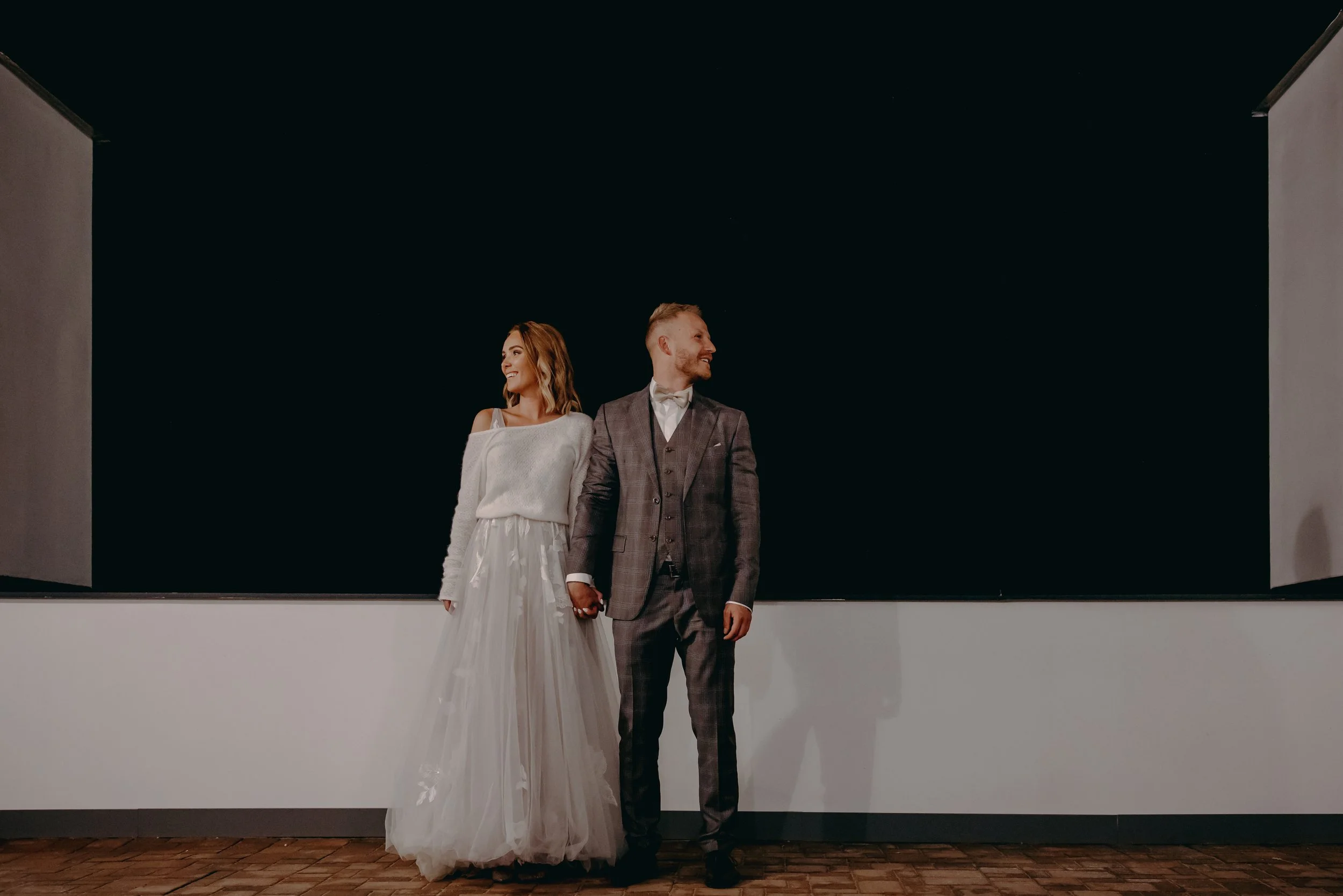 A bride and groom holding hands, standing outdoors at night against a black background, smiling and looking at each other.