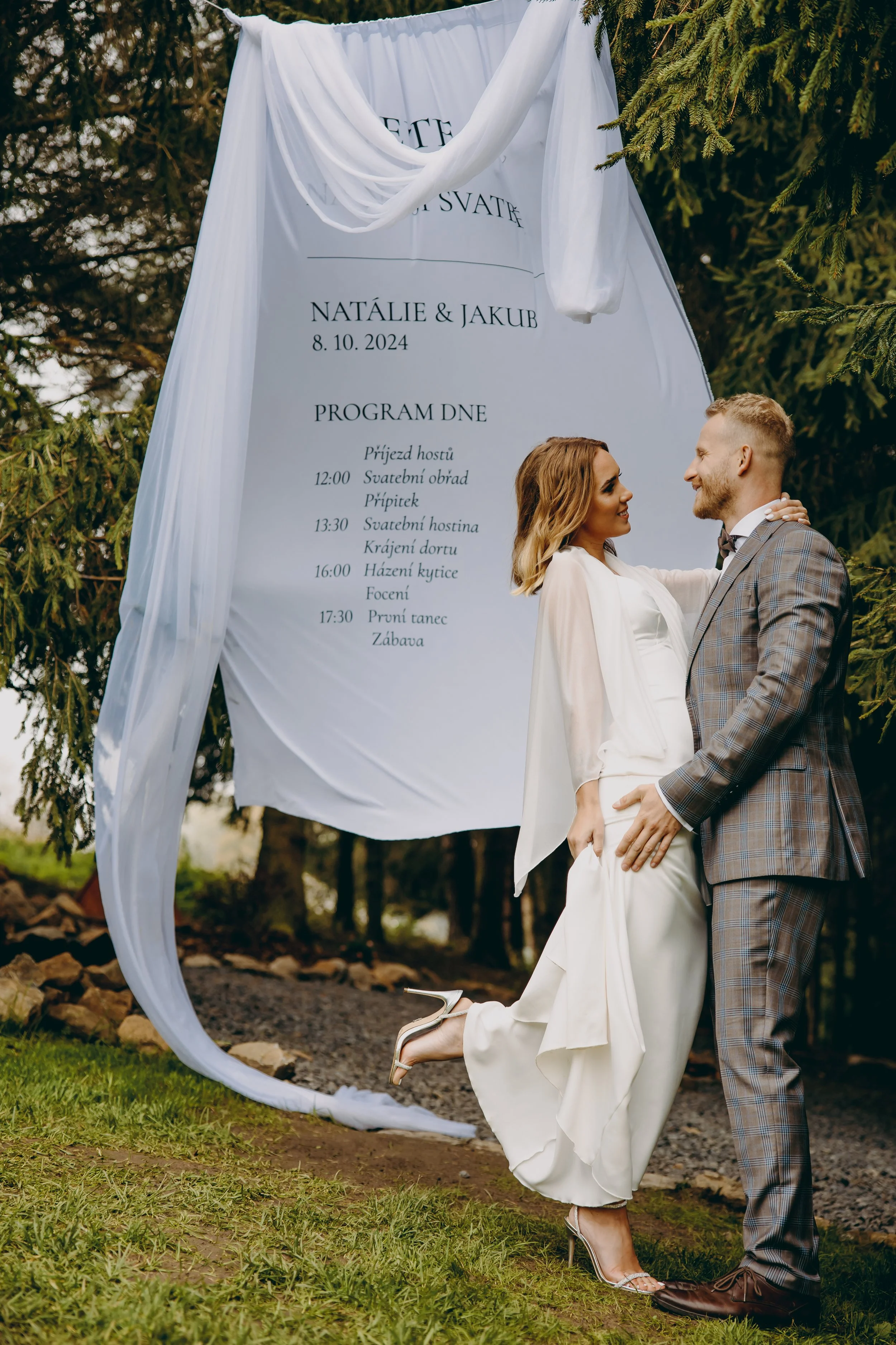 A bride and groom standing outdoors in front of a wedding schedule board. The bride is wearing a white dress and high heels, the groom is in a checkered suit. They are smiling and holding each other.
