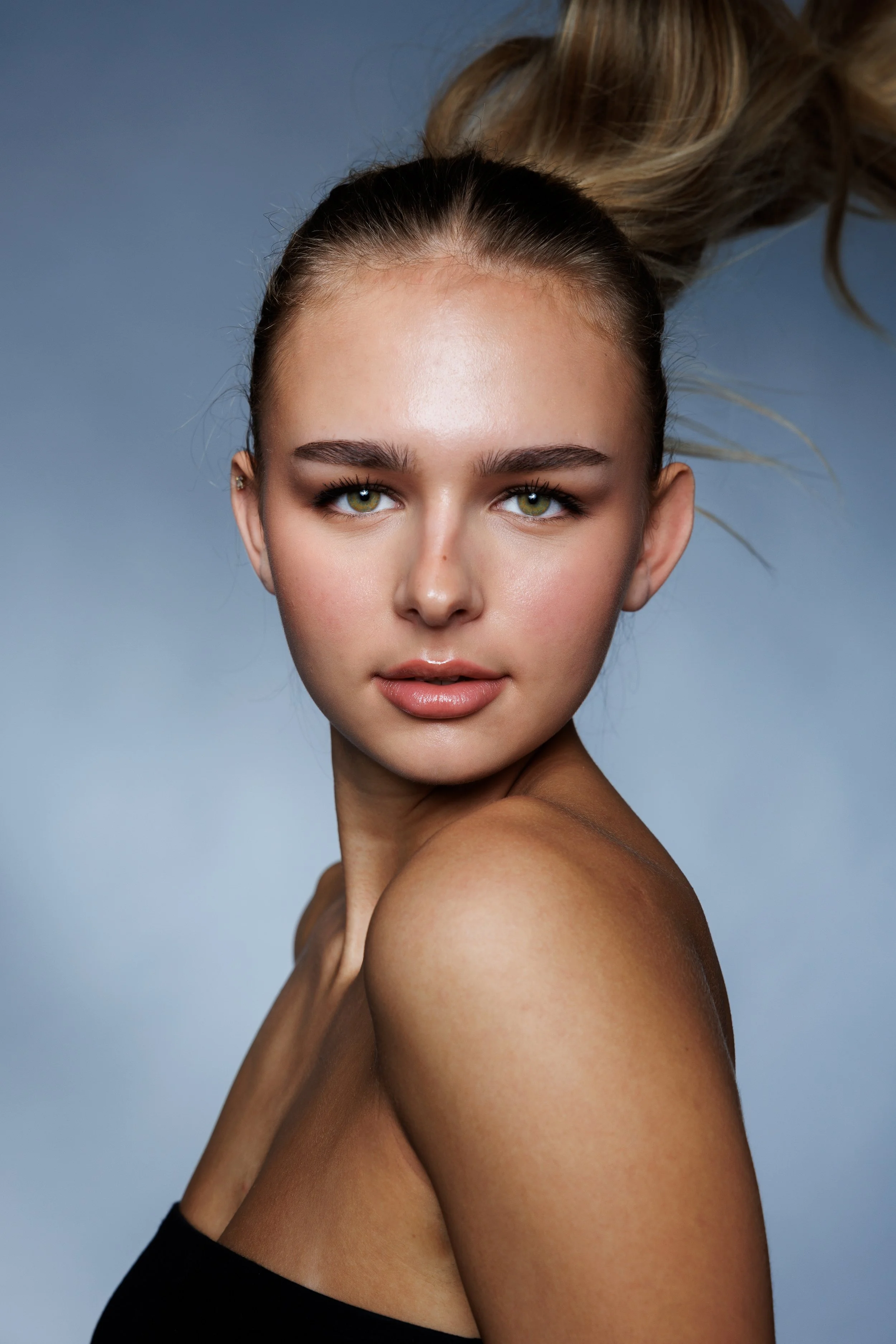 A young woman with green eyes and light skin posing with her head slightly tilted, shoulder exposed, and her hair tied up. She has minimal makeup and a confident expression against a gray background.