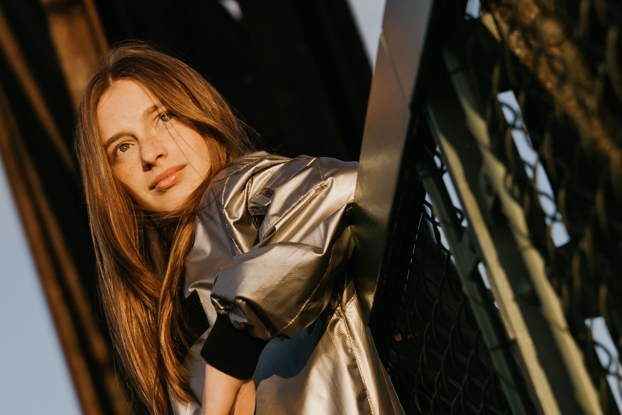 Young woman with red hair and freckles leaning against a black fence outdoors at sunset, wearing a shiny jacket.