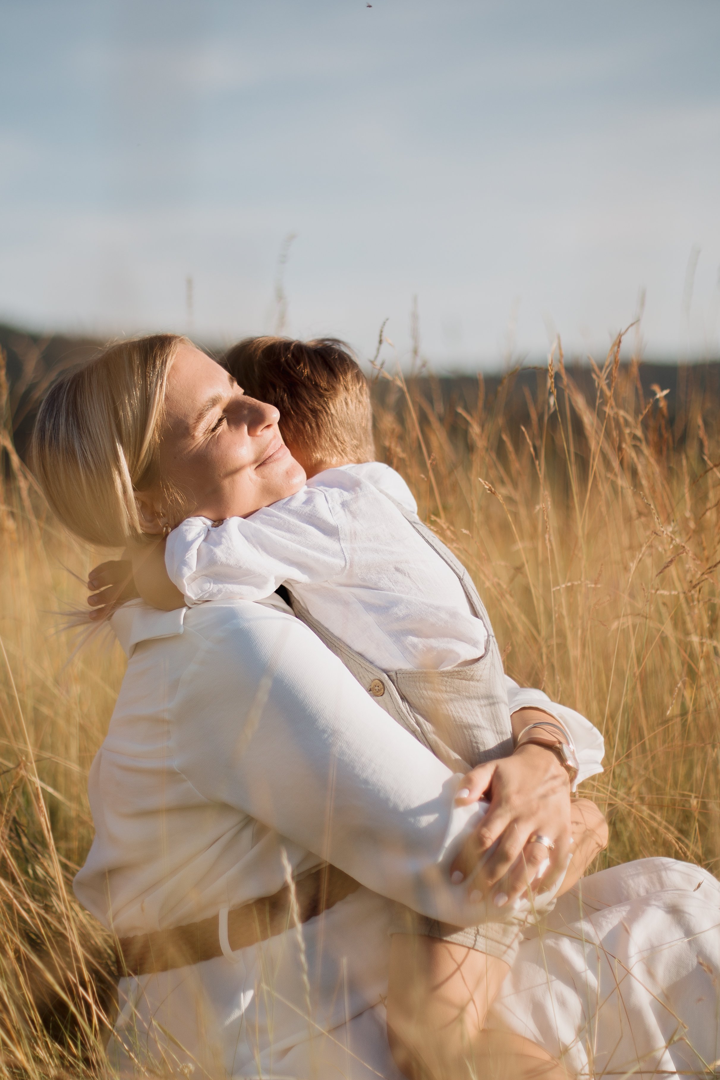 A woman with blonde hair and a child hugging in a wheat field on a sunny day.