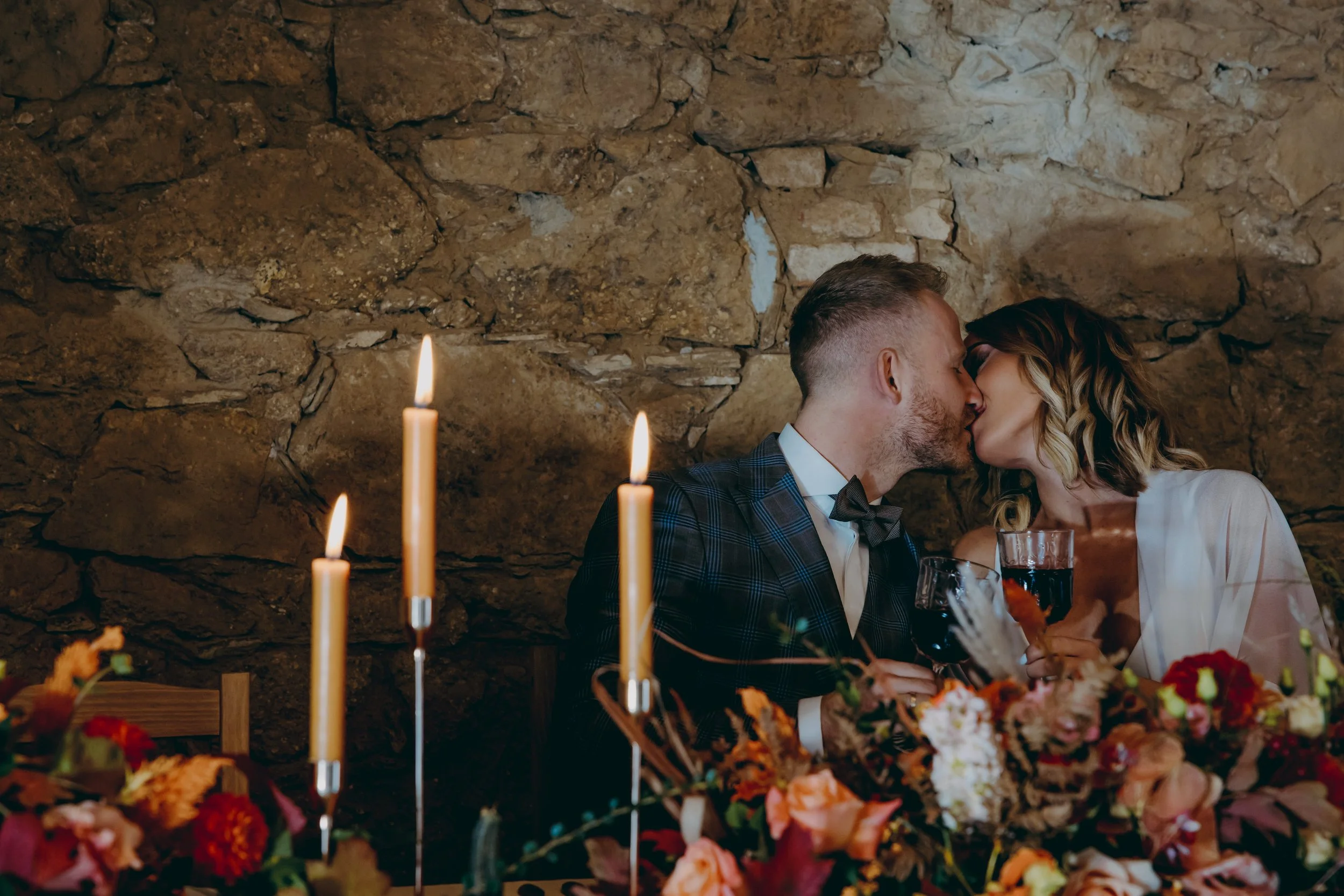 A couple sharing a kiss at a romantic dinner with lit candles and floral decorations.