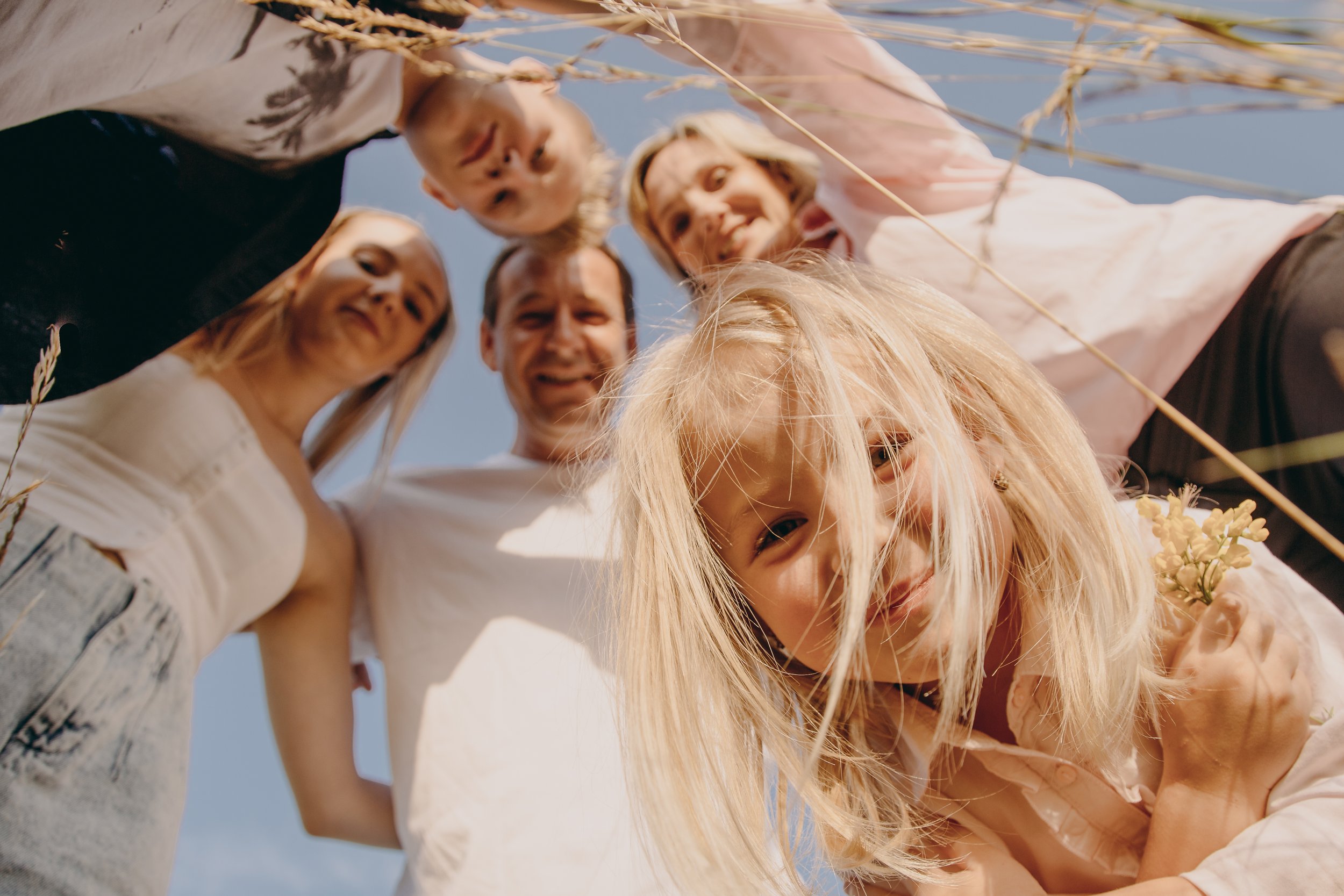 Group of six smiling people, including children and adults, looking down at the camera outdoors on a sunny day, with blue sky and dry grass around.