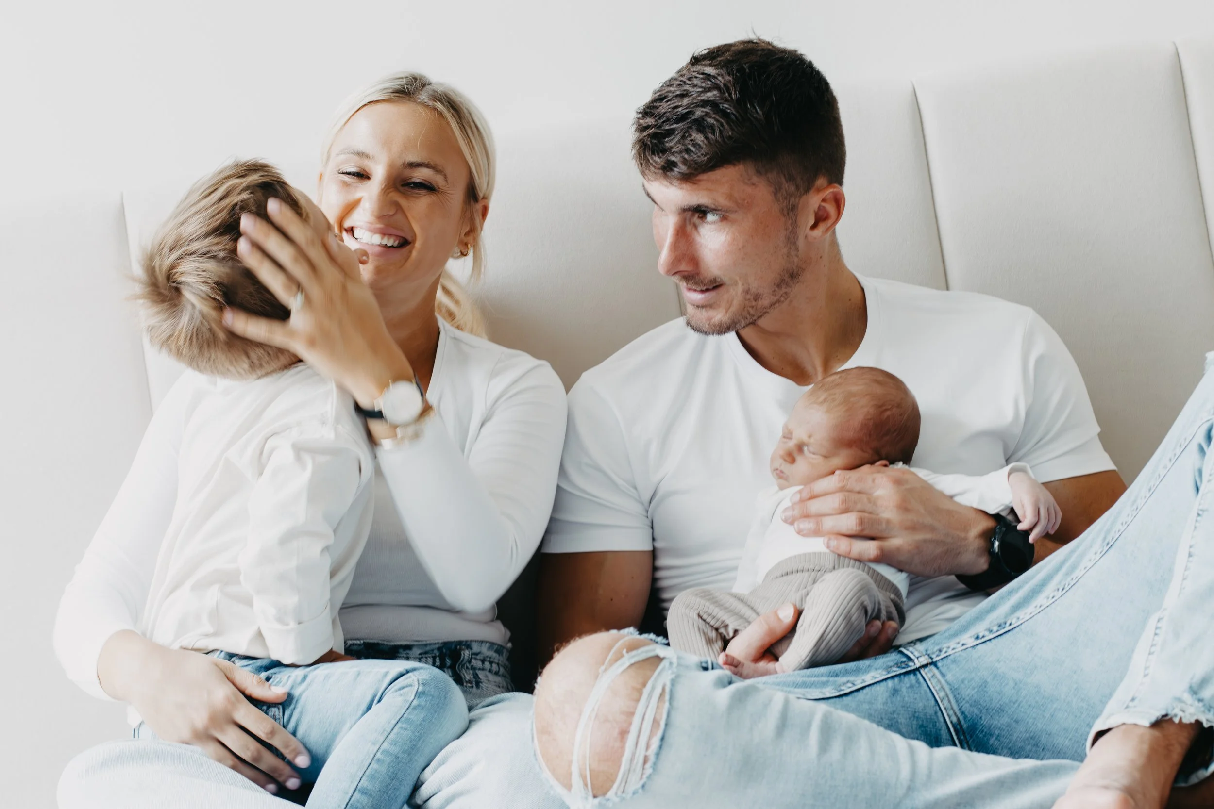A family sitting on a white bed. The mother, smiling, is holding a young child's head. The father, holding a newborn, looks at the child with affection.