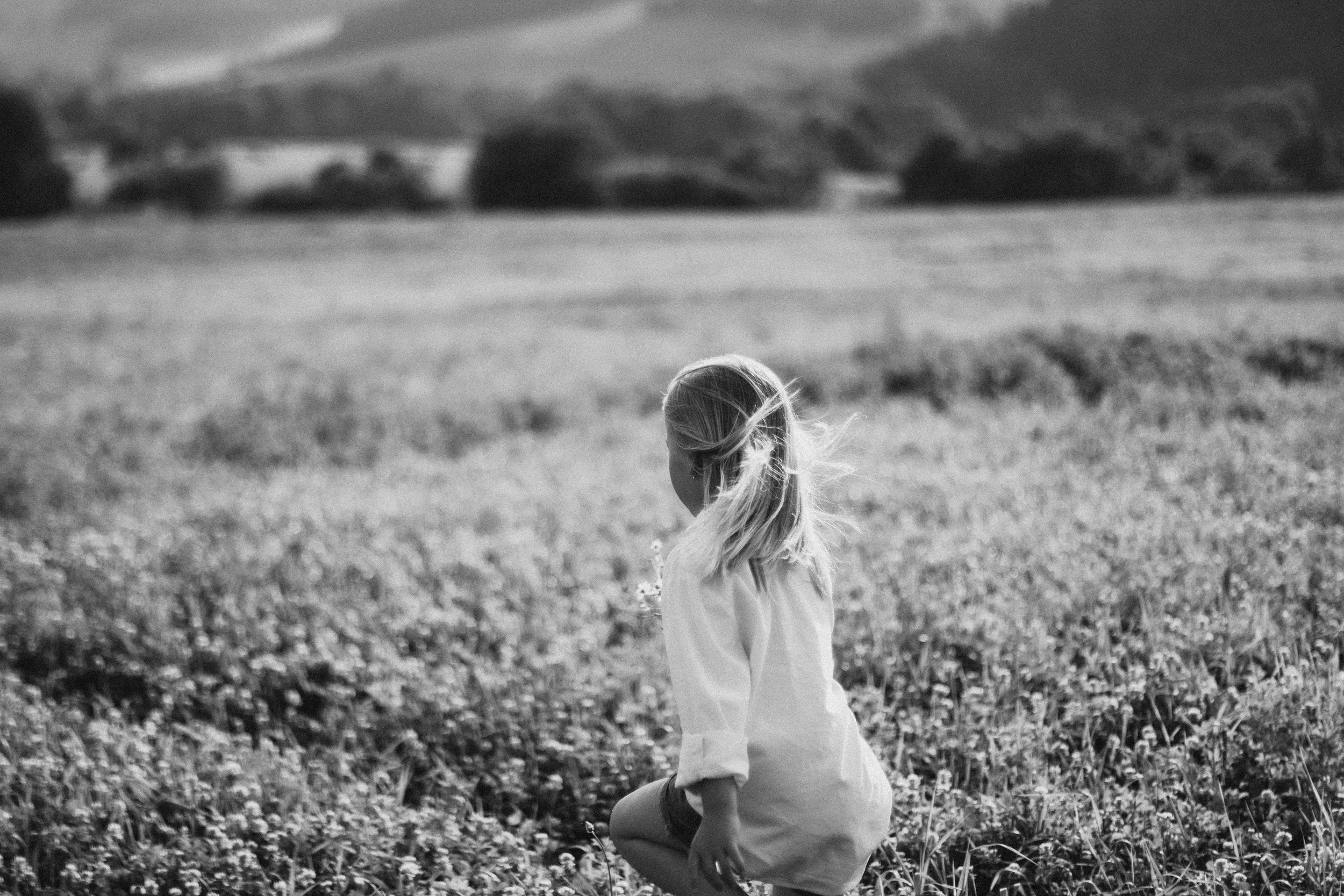 A young girl with long hair kneeling in a field of flowers, facing away from the camera, with mountains in the background.