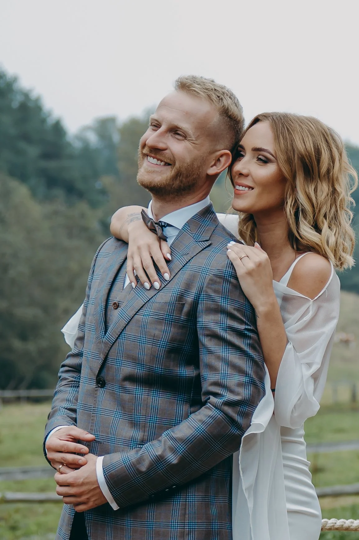 A happy couple embracing outdoors, with the woman behind the man, both smiling and looking content.