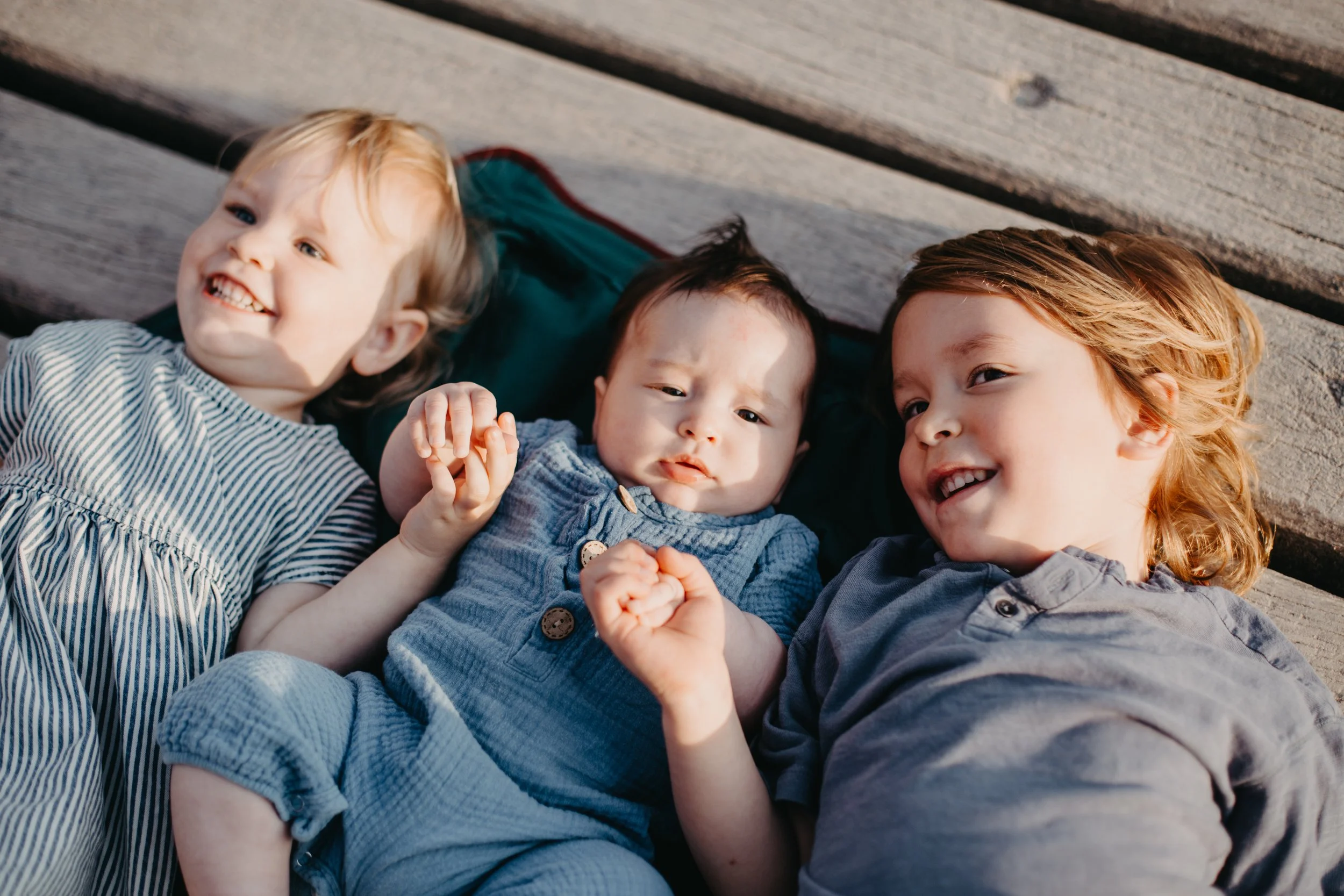 Three children lying on wooden planks, smiling and holding hands.