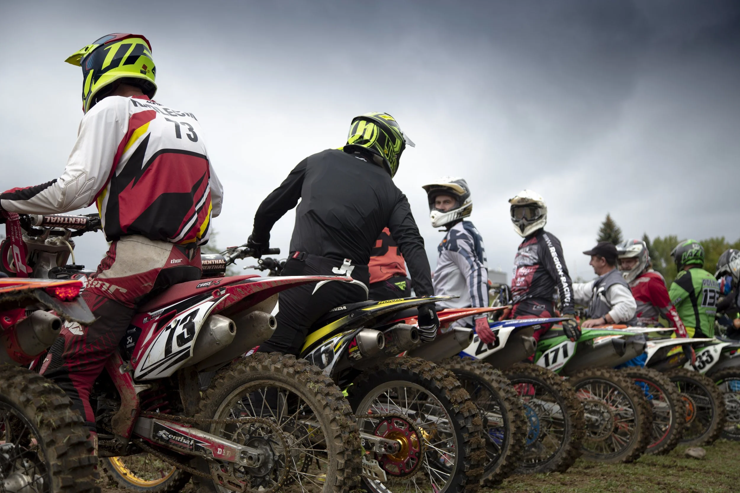 Motocross racers in gear and helmets lined up on dirt bikes before a race under an overcast sky.