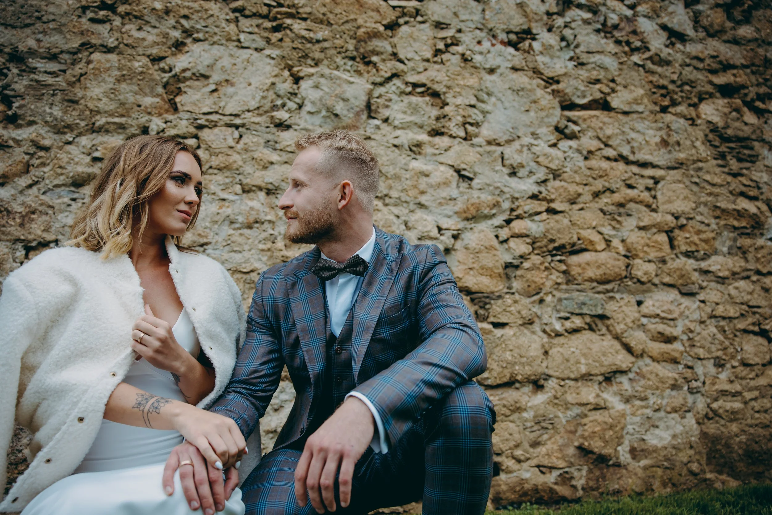 A man and woman sitting close together in front of a stone wall, gazing into each other's eyes, dressed in formal attire with the woman in a white dress and the man in a plaid suit.