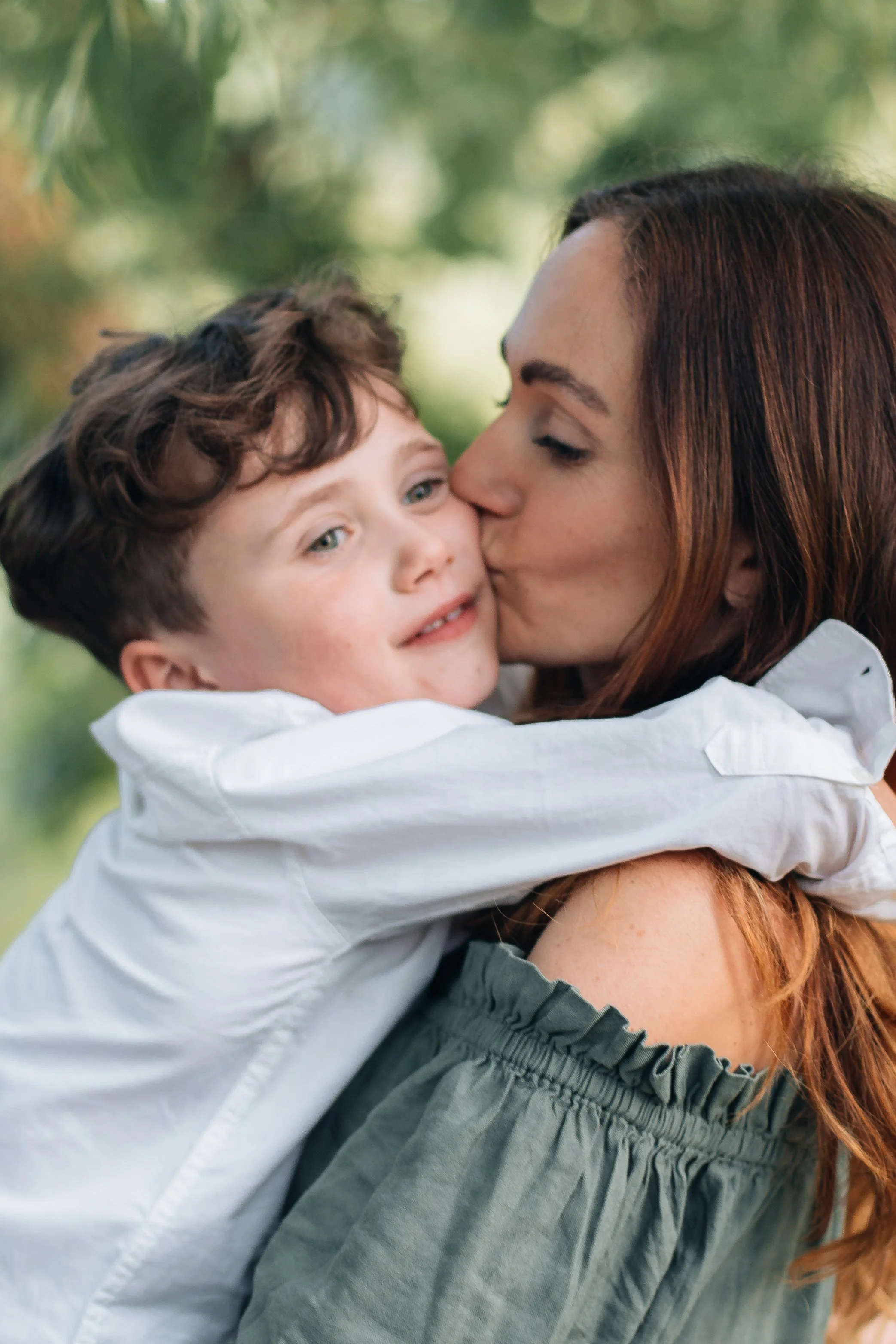 A woman with red hair kisses a young boy on the cheek as he embraces her, outdoors with blurred green trees in the background.