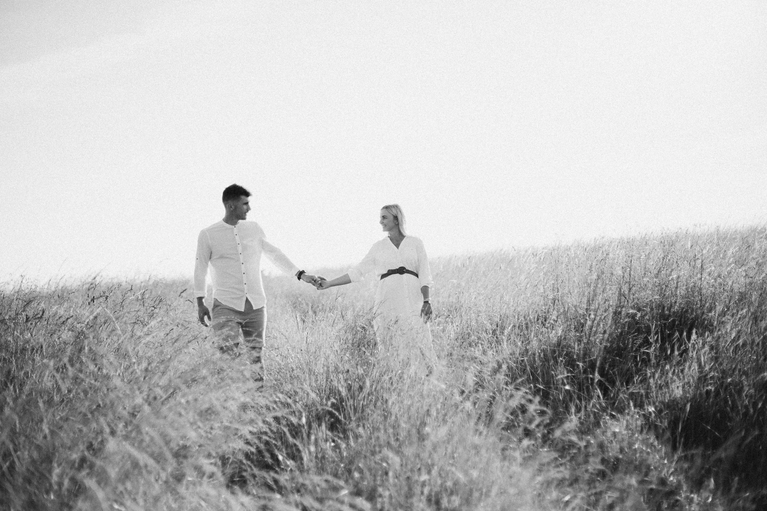 A Black and white photo of a couple holding hands and walking through tall grass in a wide open field.