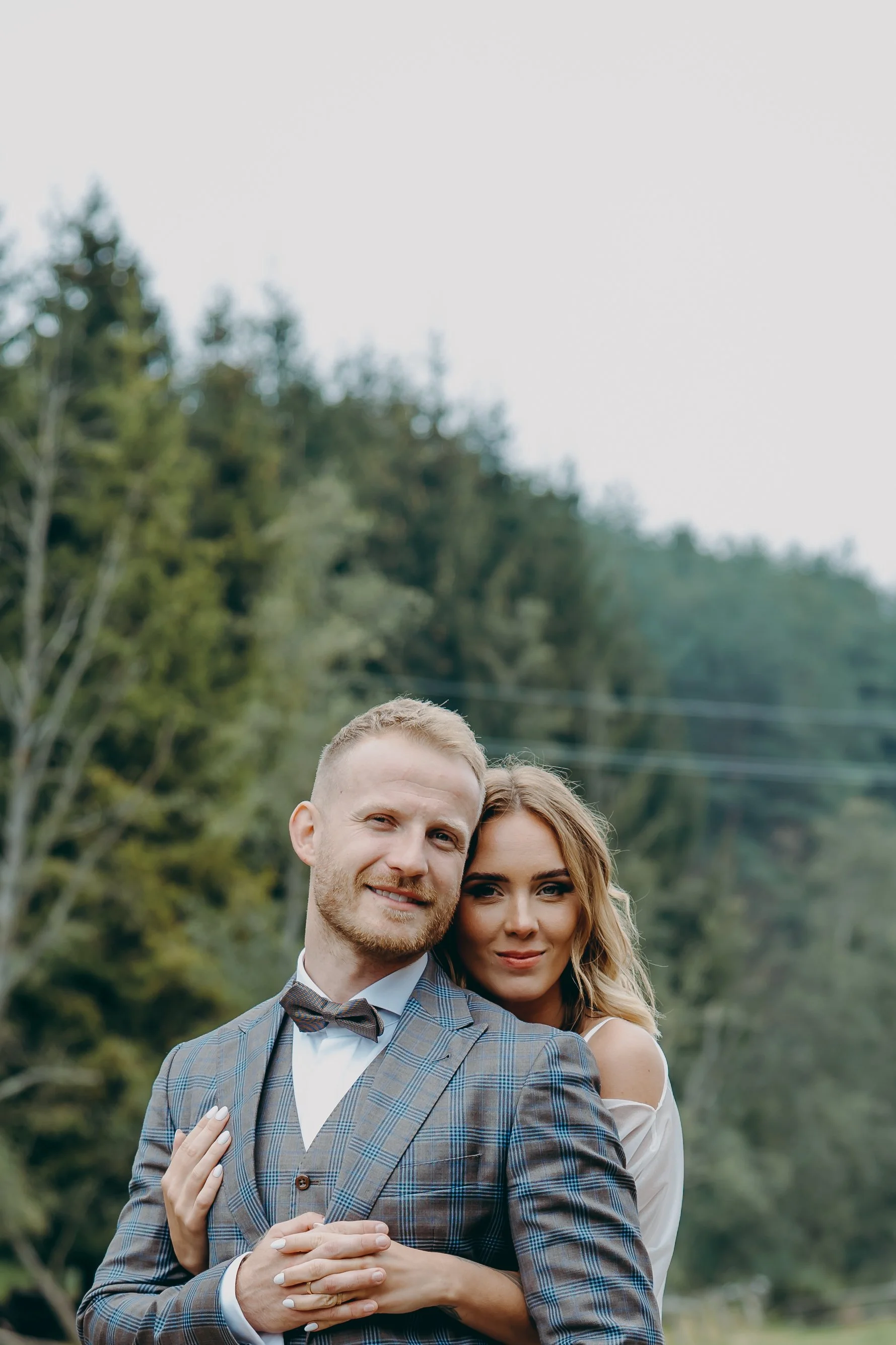 A couple dressed in formal attire embracing outdoors with trees in the background.