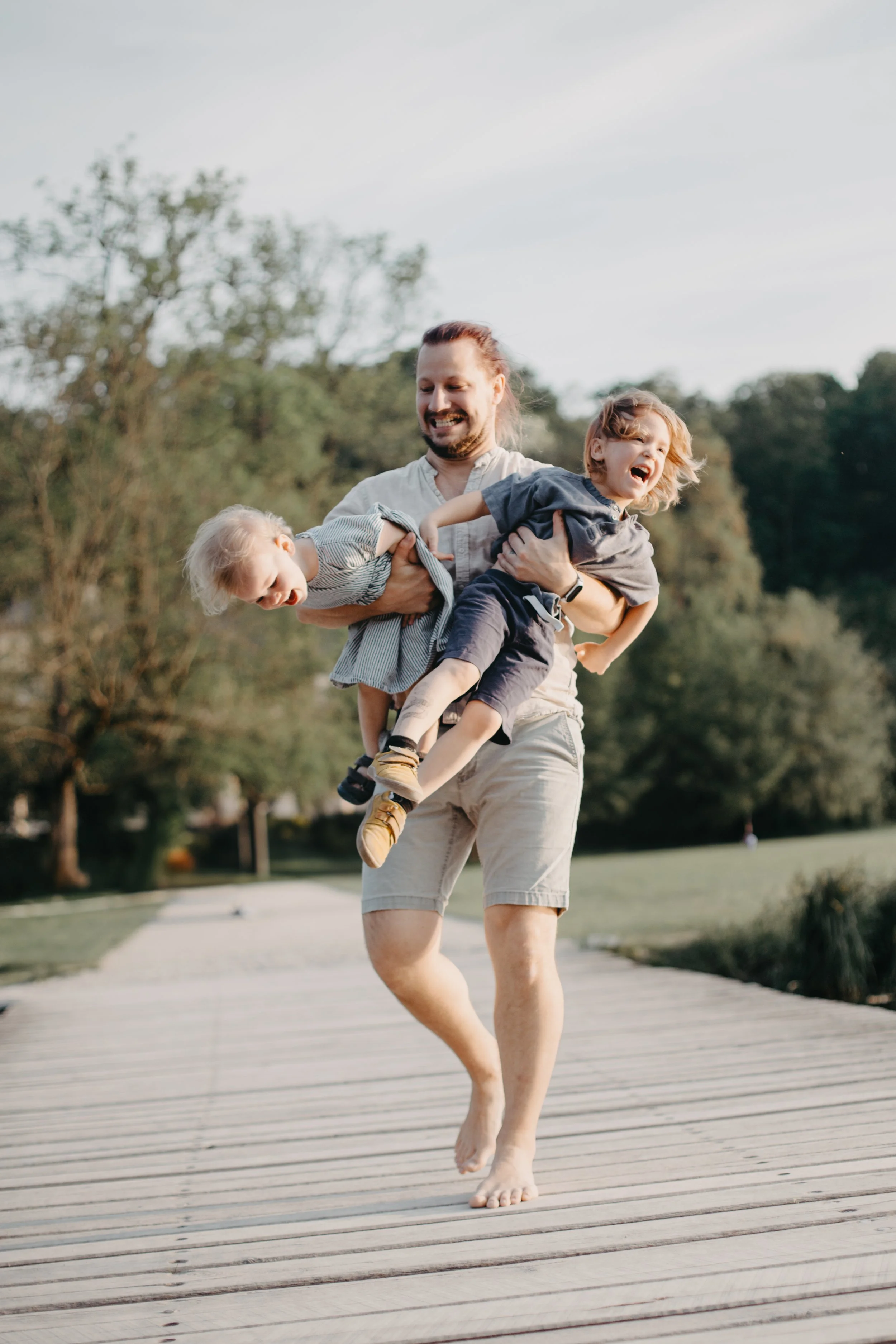 A man with long hair and a beard, wearing shorts and a light shirt, is barefoot and joyfully running on a wooden pathway while holding two young children, one girl and one boy, who are laughing and enjoying the moment outdoors with trees and sky in the background.