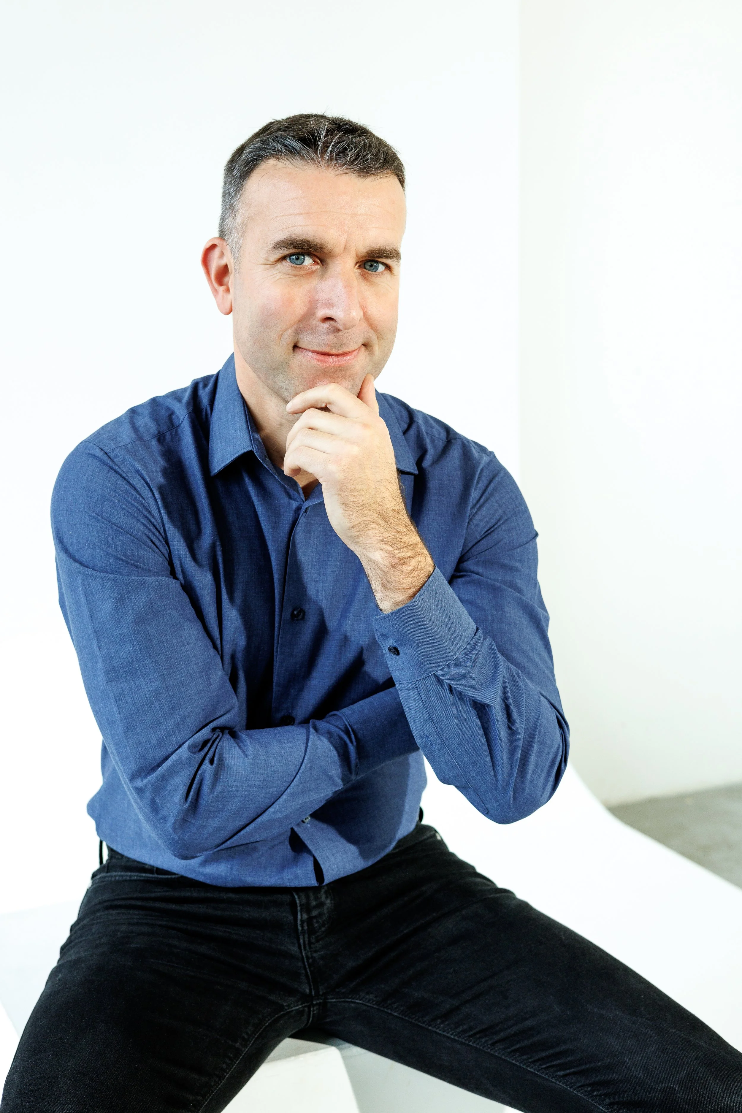A man with short dark hair, blue eyes, wearing a blue button-up shirt and black pants, sitting and posing confidently with his hand on his chin against a plain white background.