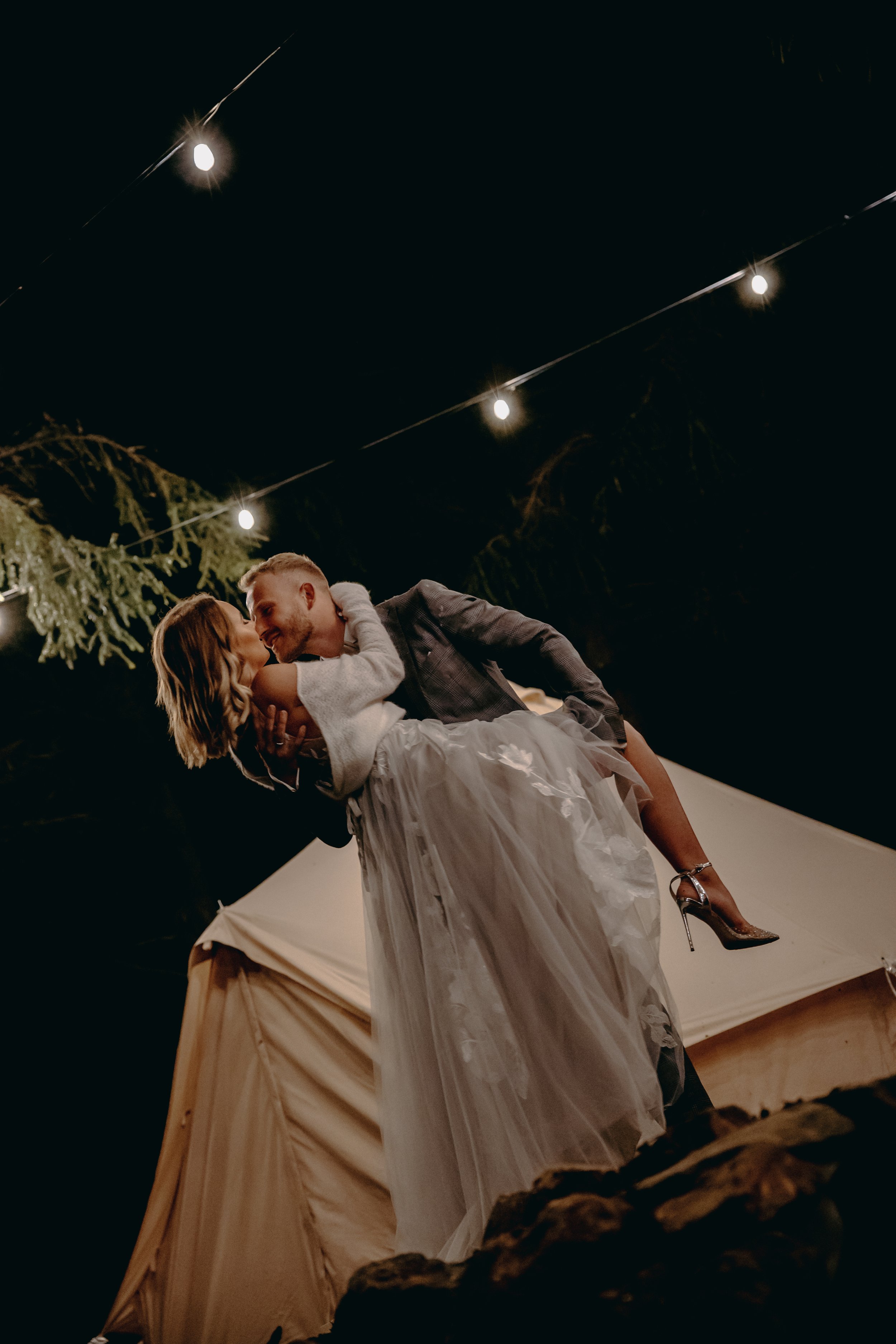 A couple dancing at an outdoor nighttime event, with string lights overhead and a tent in the background, the man is lifting the woman.