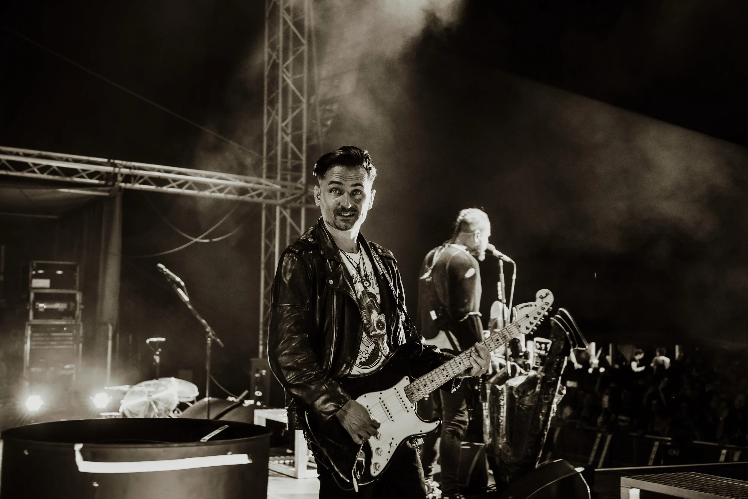 Black and white photo of a musician holding an electric guitar on stage during a concert at night, with another musician singing into a microphone in the background, stage equipment, and lighting.