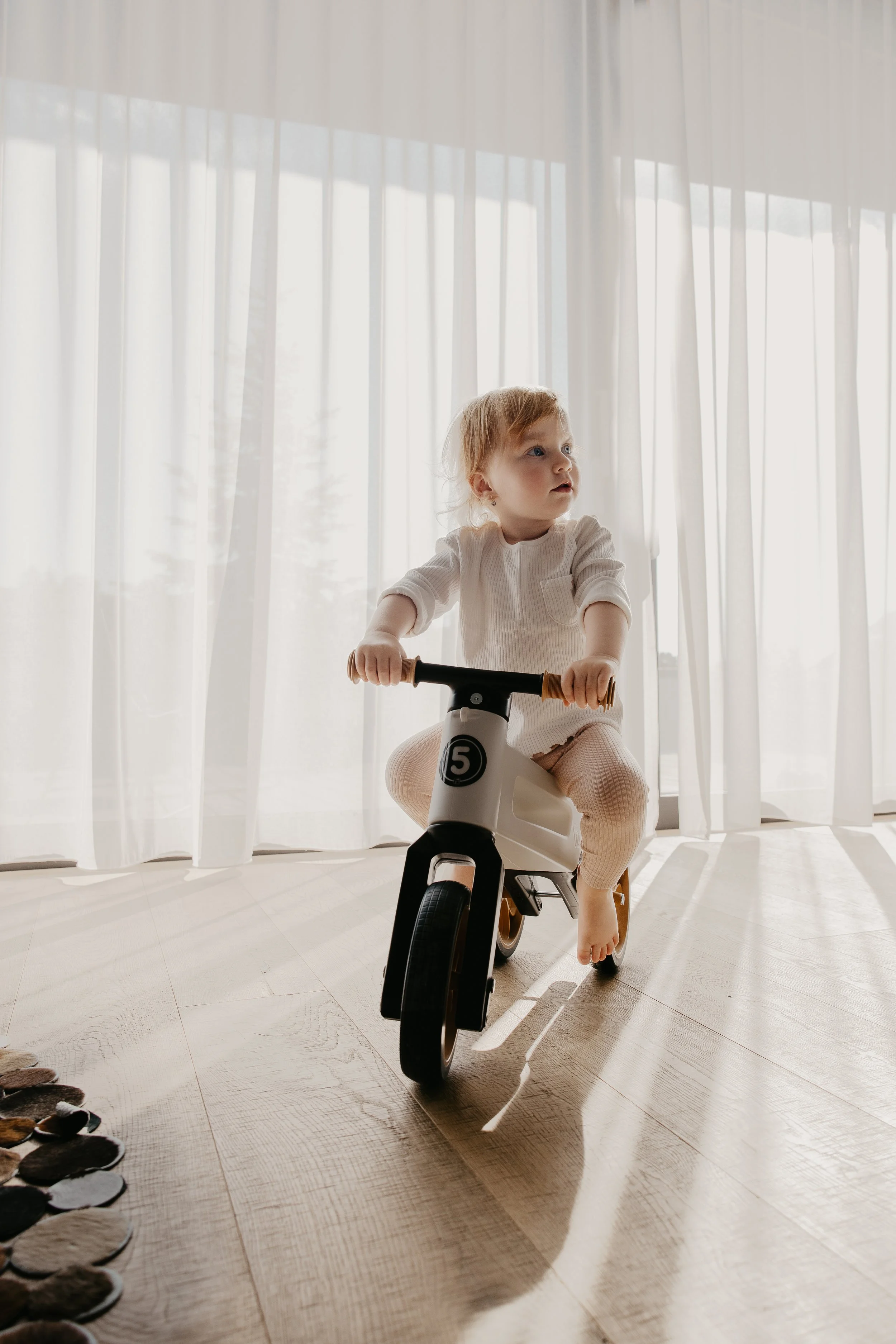 A young child riding a balance bike indoors near large windows with sheer curtains.