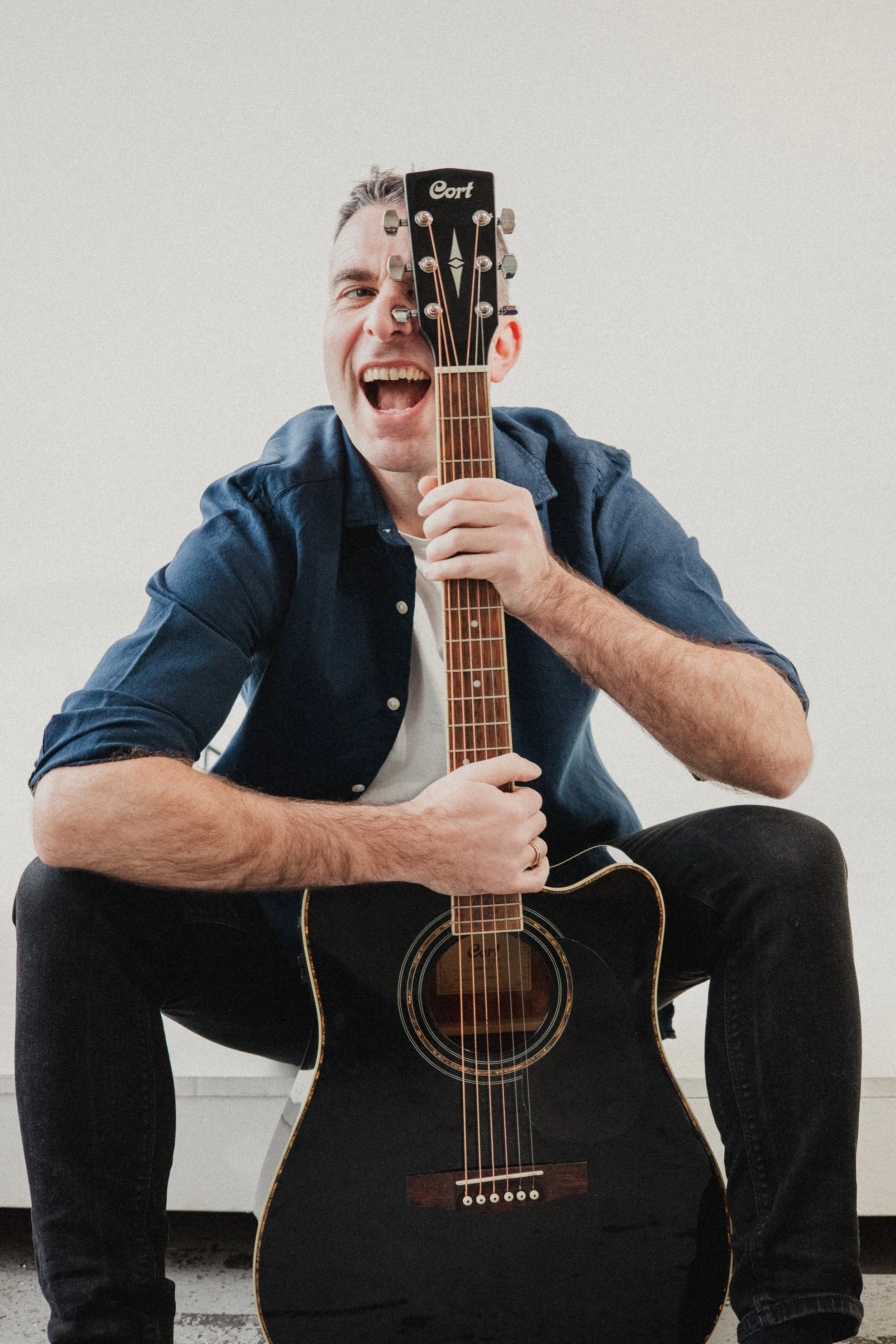 A man sitting on the floor, smiling and holding an acoustic guitar vertically in front of him, with the guitar's headstock covering one eye.