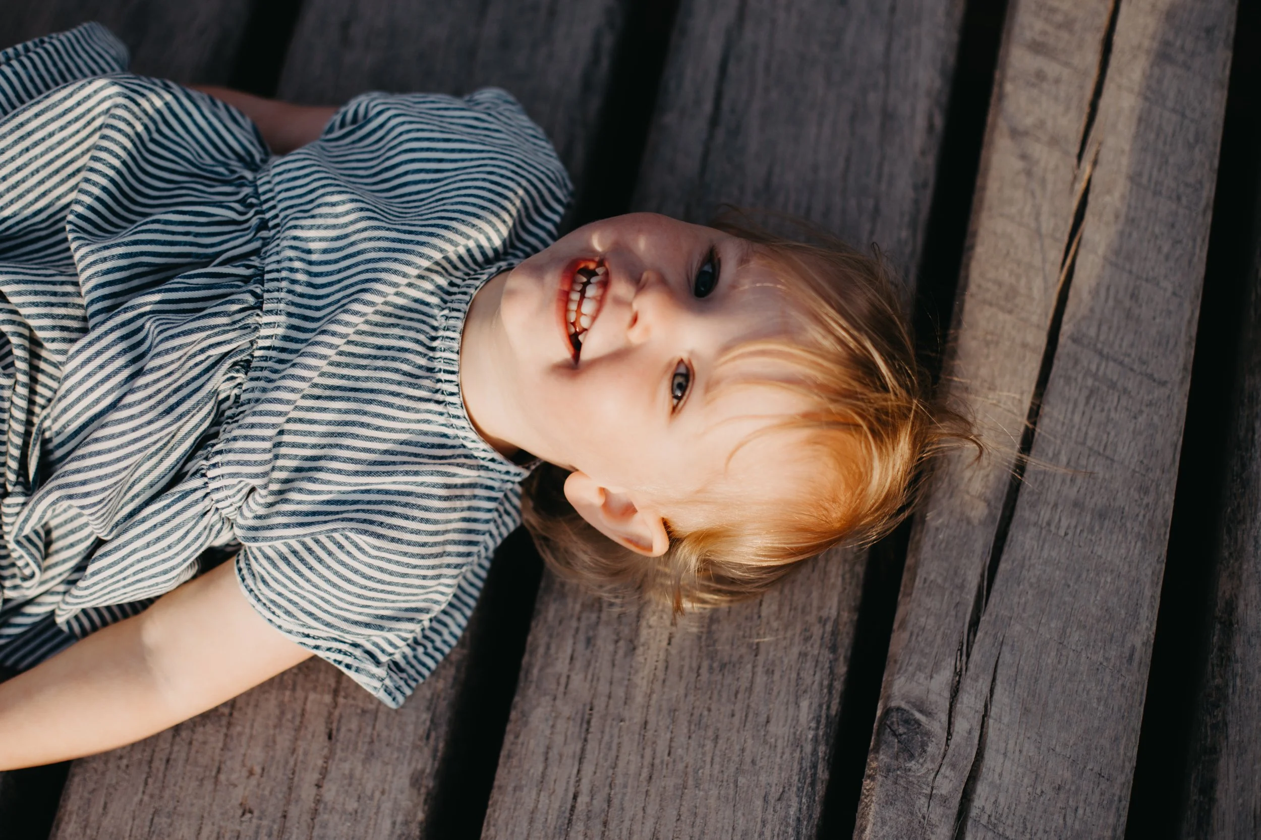 A young girl with blonde hair lying on a wooden surface, smiling and looking up.