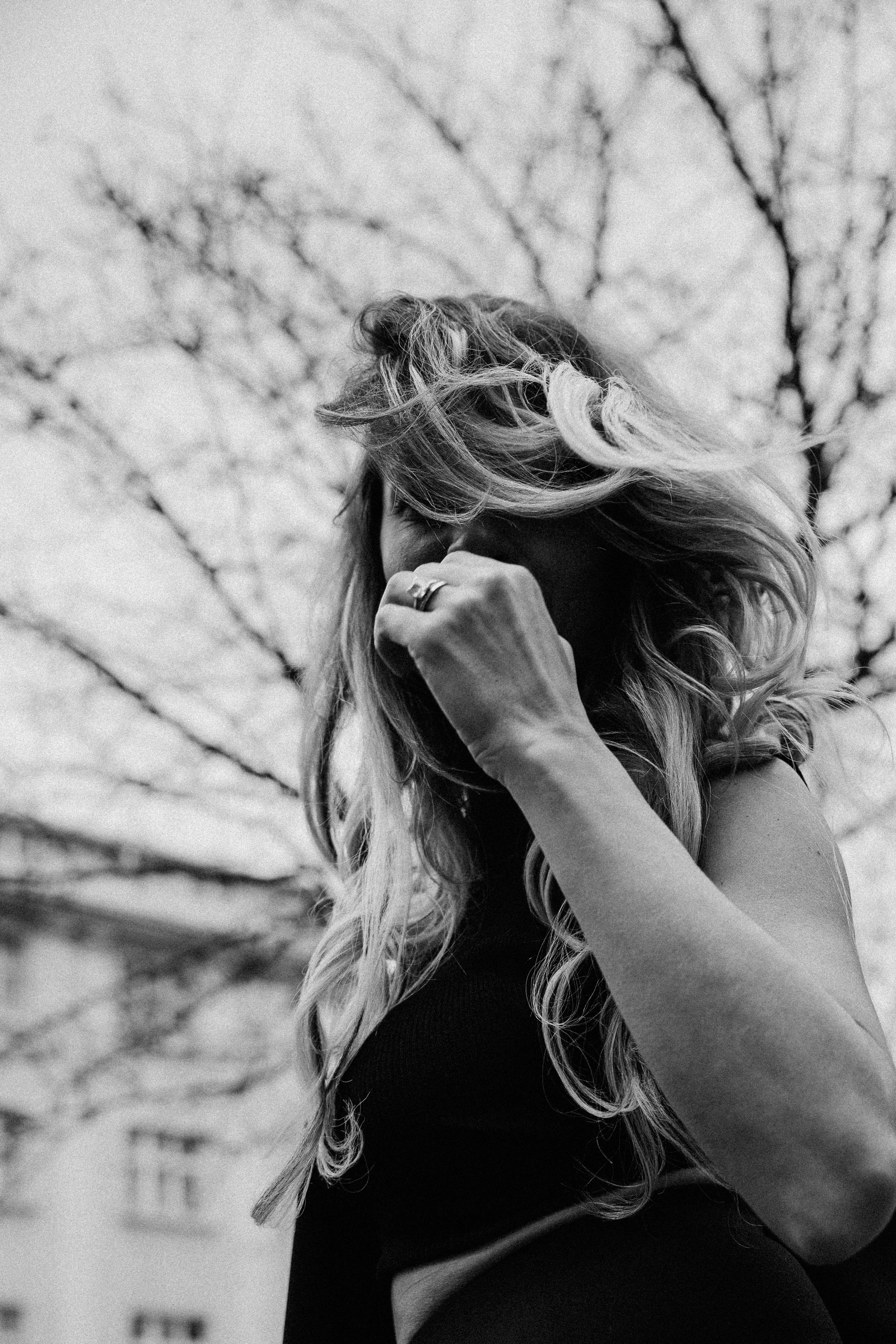 Black and white photo of a woman with long, wavy hair covering part of her face, standing outdoors with tree branches in the background. She is wearing a sleeveless top and has her hand near her face.