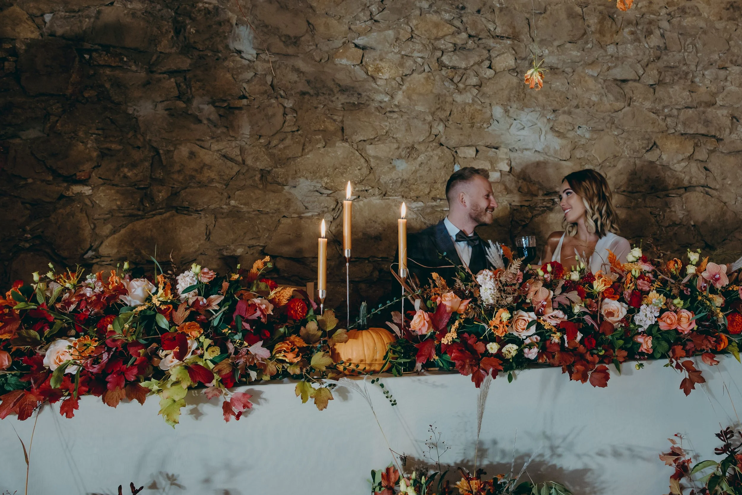 A couple at a wedding reception sitting at a table decorated with autumn-themed floral arrangements, candles, and a pumpkin, gazing at each other and smiling.