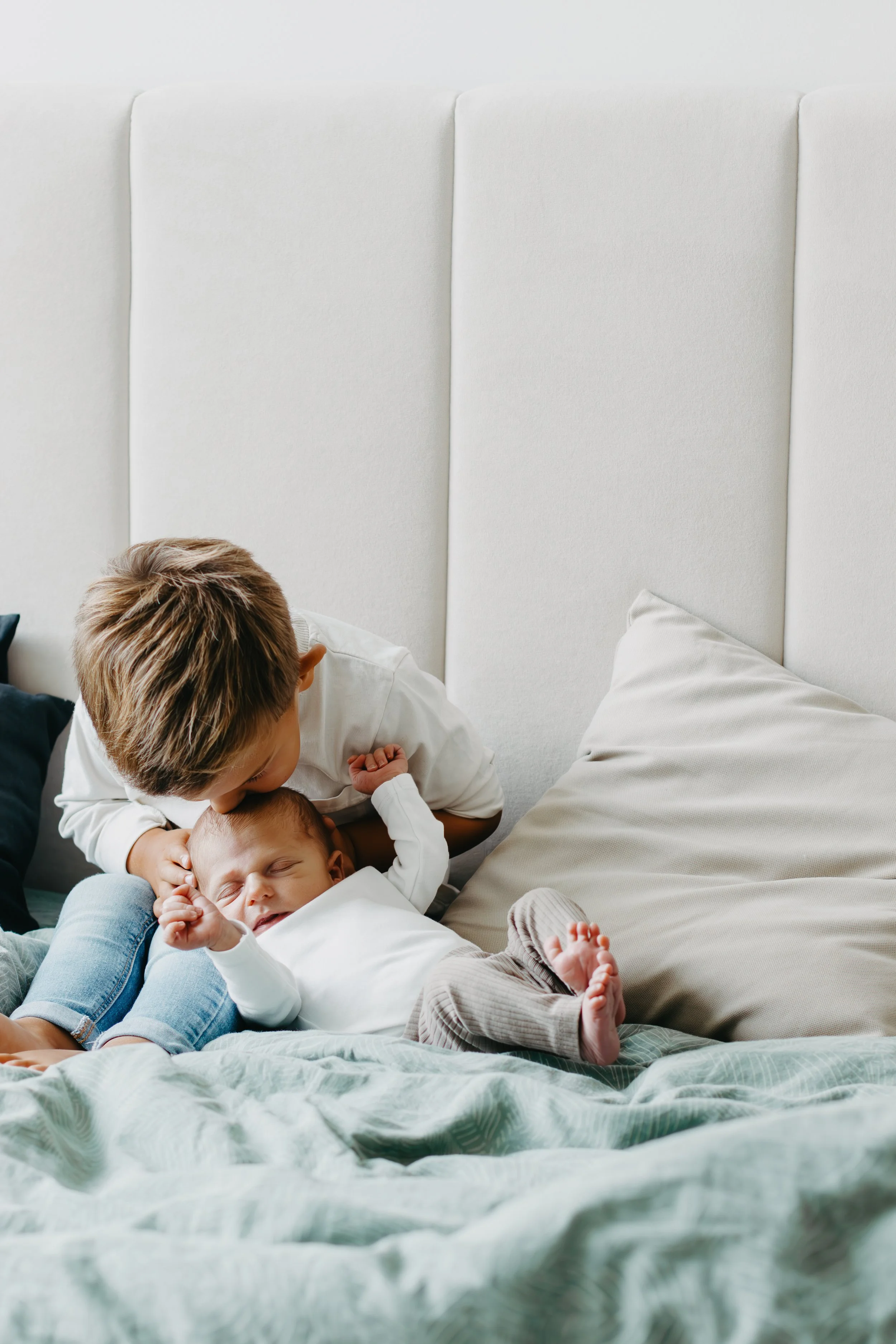 A young boy giving a kiss on the forehead to a sleeping baby on a bed, with the boy leaning over the baby.