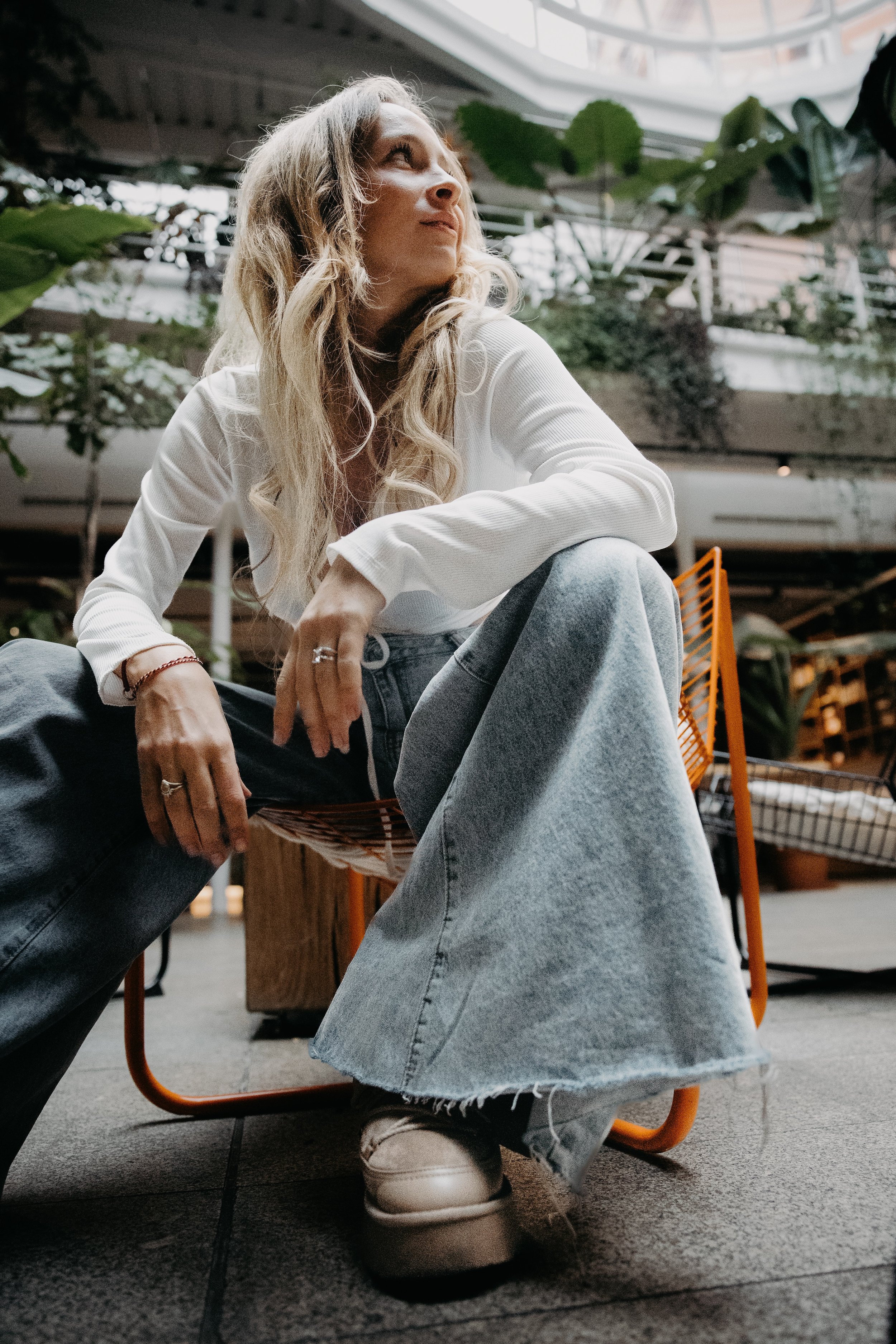 A woman with long blonde hair wearing a white long-sleeve shirt, light-wash jeans, and beige sneakers sitting on a modern orange chair in an indoor setting with plants and a glass ceiling.